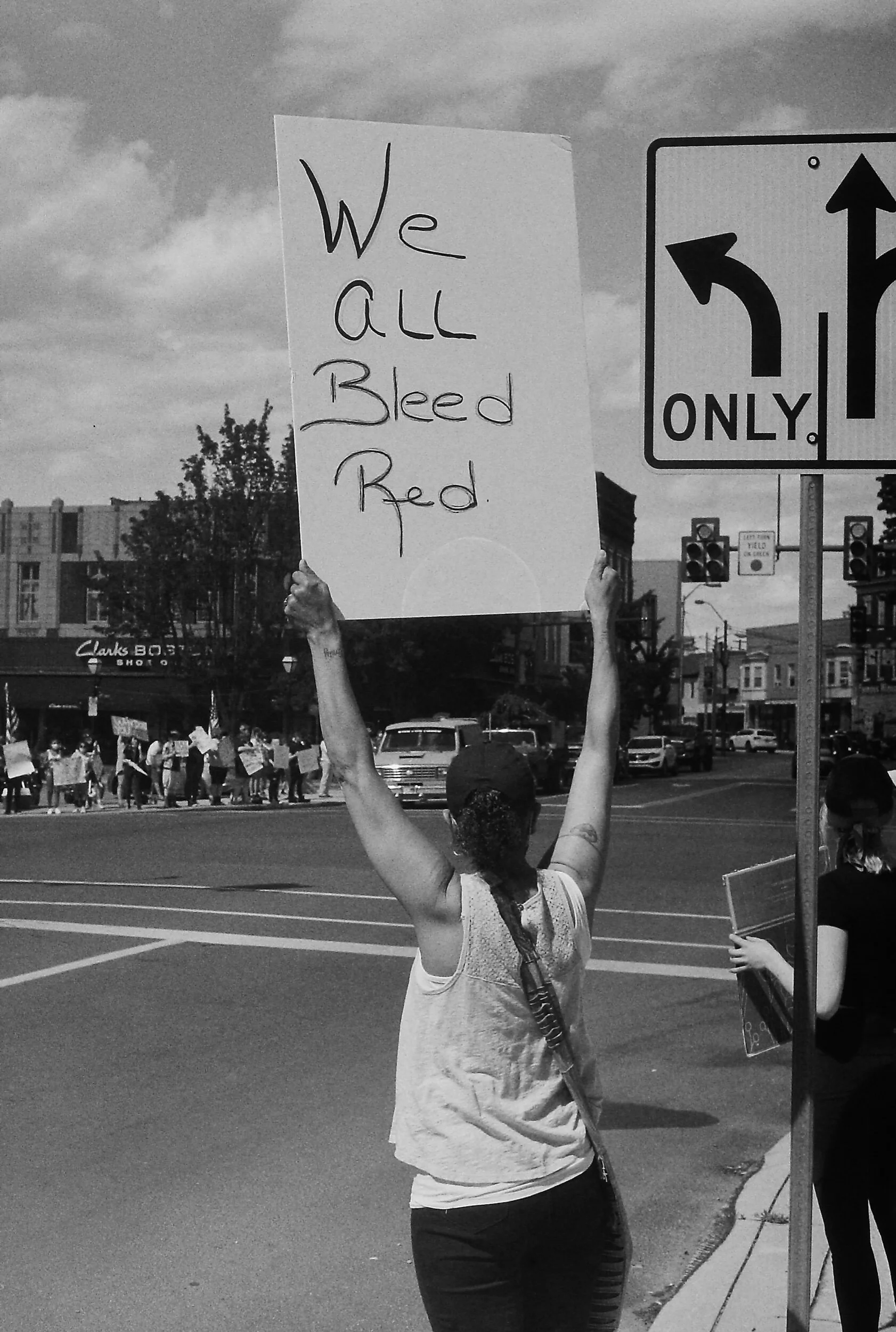 A person holds a sign that reads "We All Bleed Red" at a street protest.