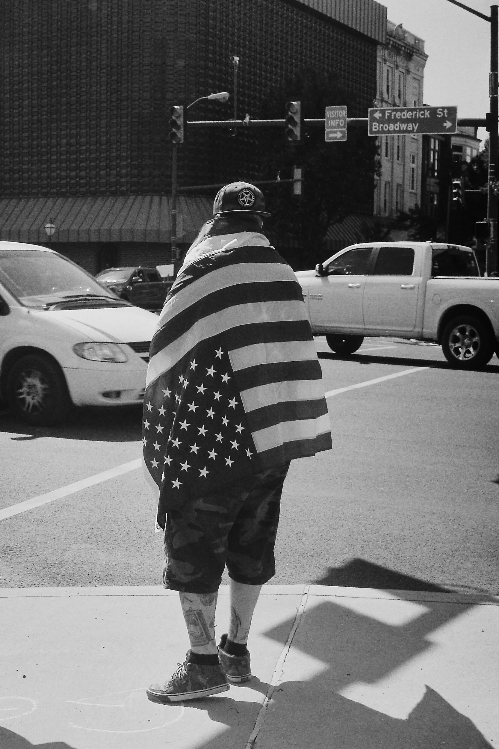 Black and white photo of a person standing at a street intersection, wearing an American flag as a cape. Cars and street signs are visible in the background.