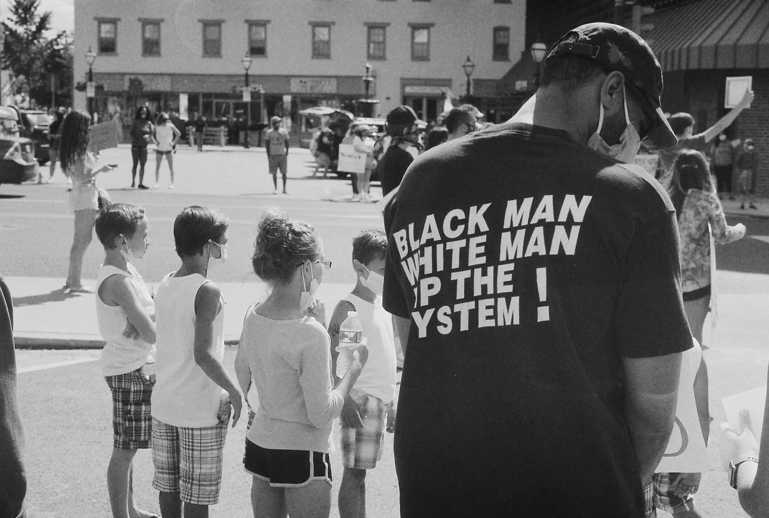 A black and white photo of a group of people, some children wearing masks, at a demonstration. A man in the foreground wears a T-shirt with the text "Black man, white man up the system!" in large letters. Background shows buildings and more people ho