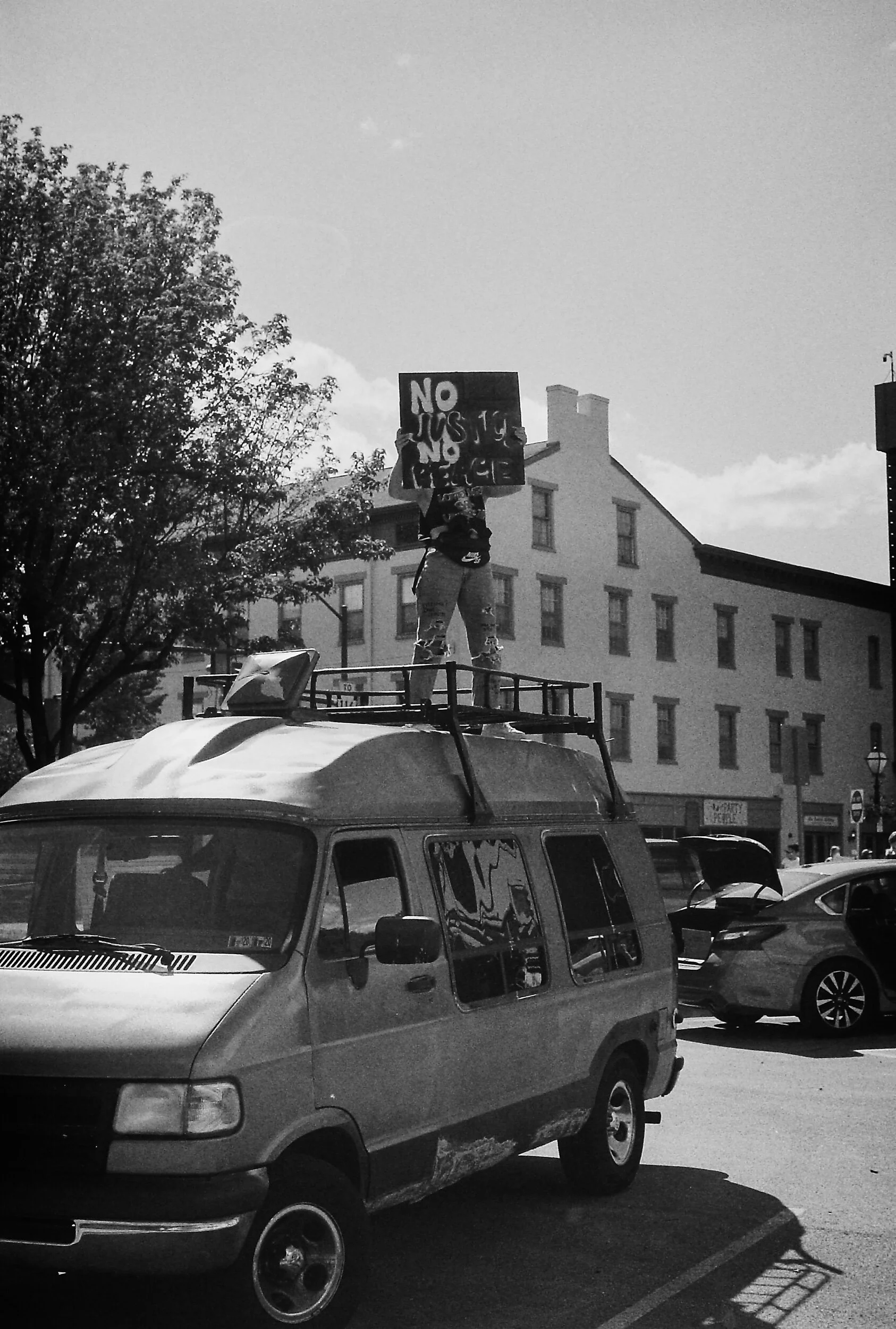 Person standing on top of a van holding a protest sign reading 'No Justice No Peace'