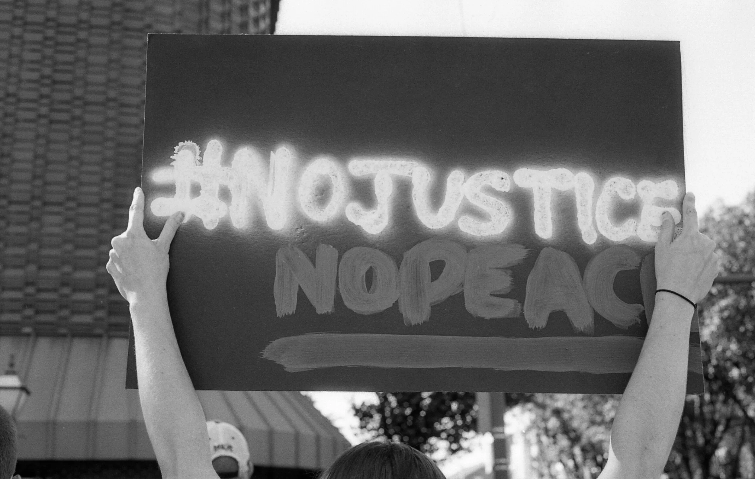 Protester holding sign with "#NoJustice NoPeace" in large letters, outdoors, in black and white photo.