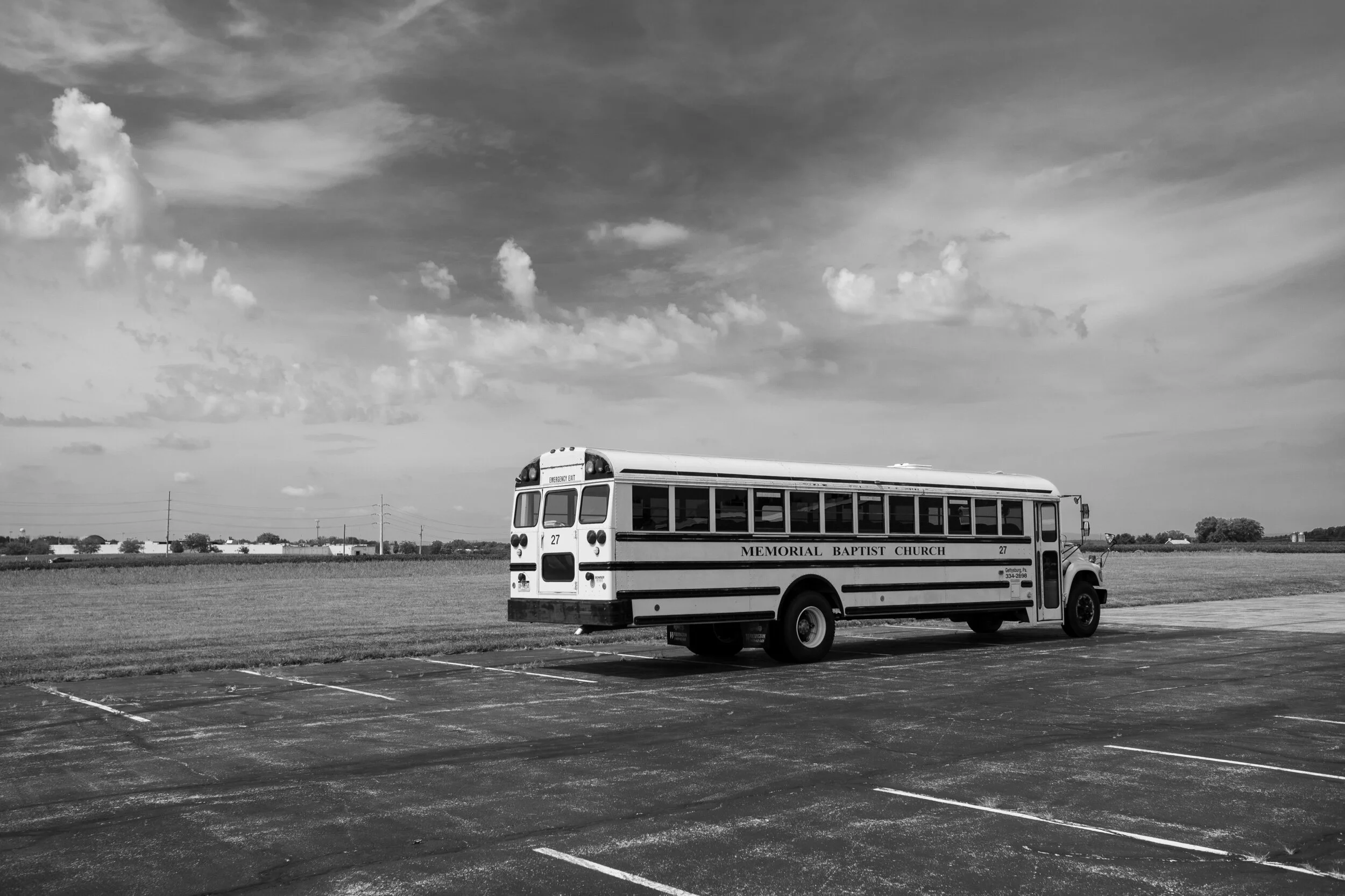 Black and white image of a parked school bus labeled 'Memorial Baptist Church' in an empty parking lot with a grassy field and cloudy sky in the background.