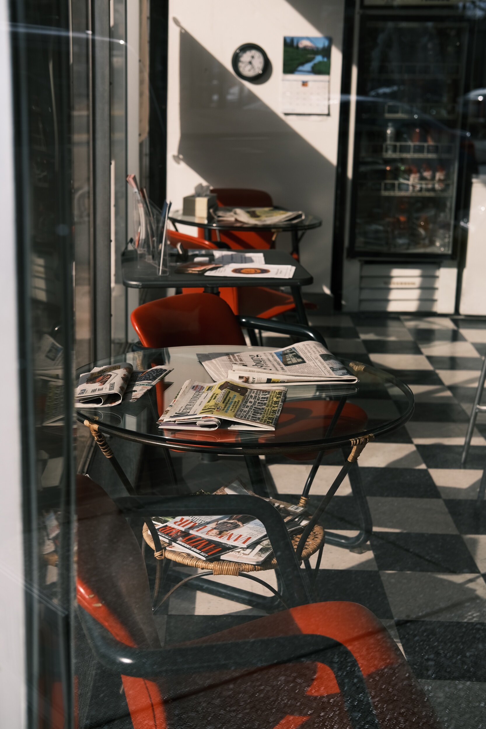 Indoor cafe seating area with glass tables, red chairs, newspapers, and magazines, black-and-white checkered floor, wall clock, and vending machine.