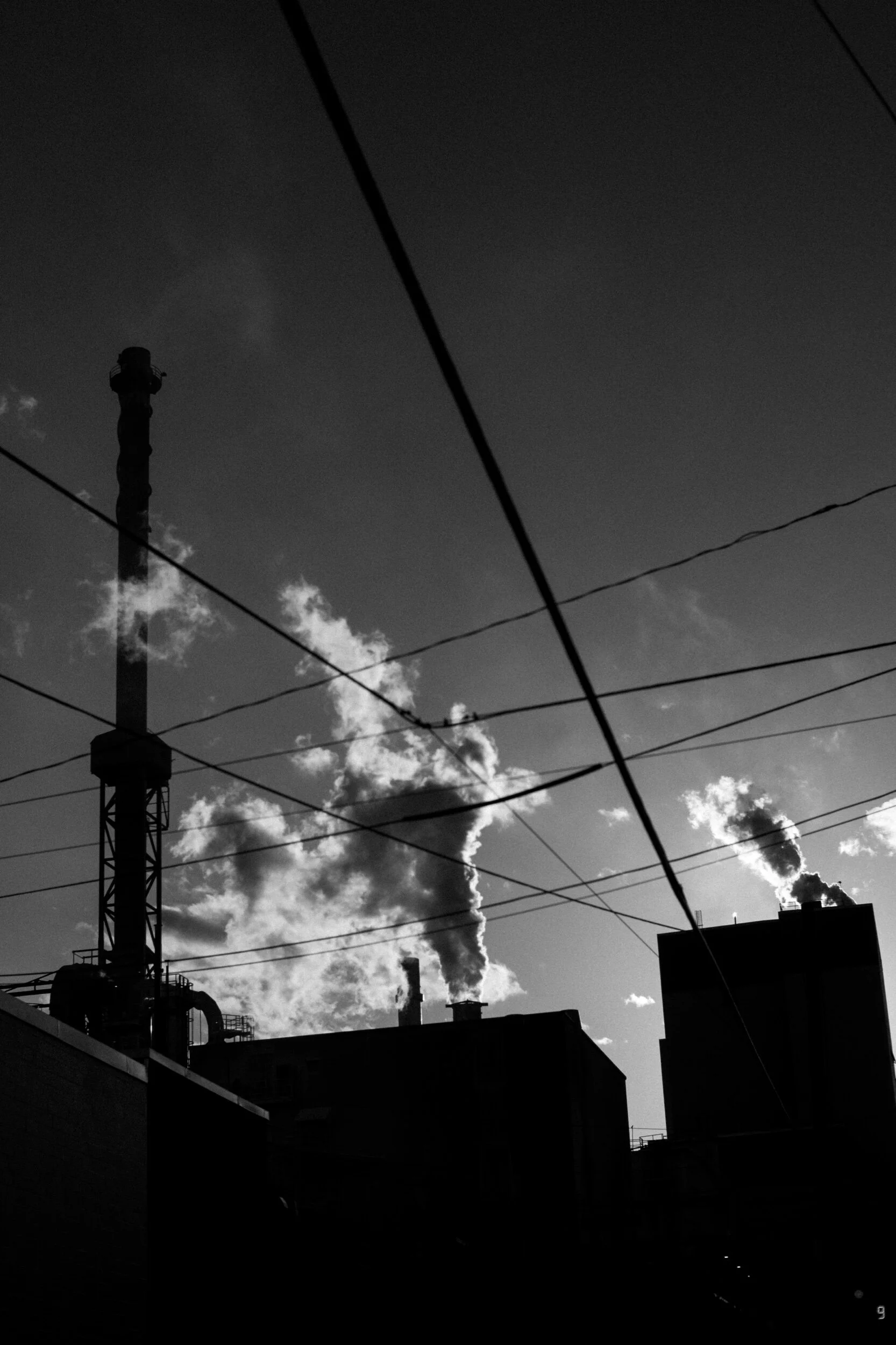 Silhouetted industrial buildings with smokestacks releasing smoke against the sky, intersected by power lines.