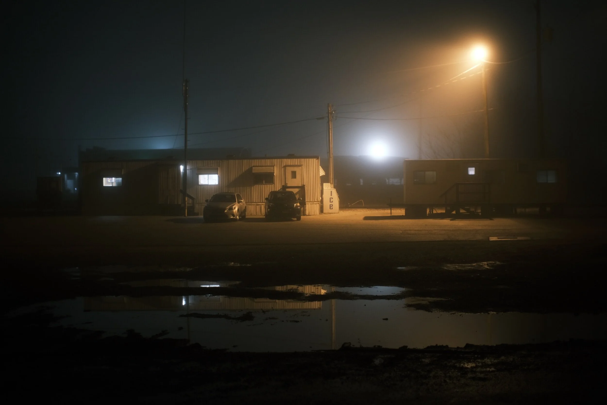 Dimly lit night scene of a foggy outdoor area with parked cars, illuminated trailers, and a large ice vending machine.