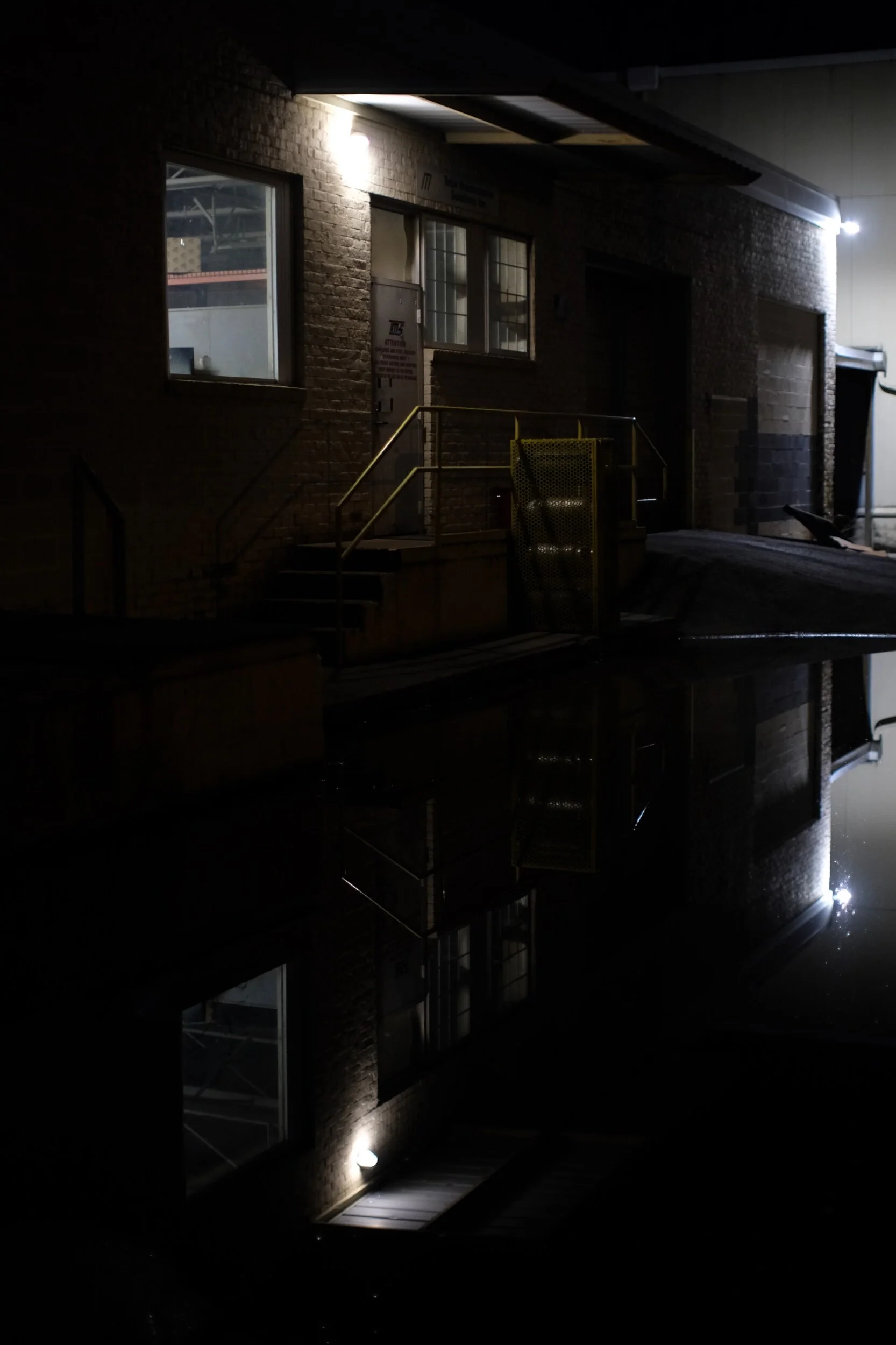 Dimly lit industrial building at night with illuminated windows and reflections in a puddle.