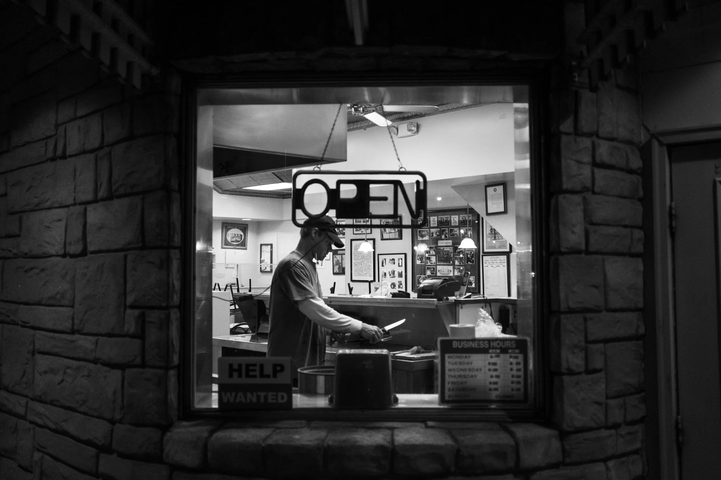 Black and white photo of a man working inside a restaurant with an 'Open' sign in the window and job advertisement for help wanted.