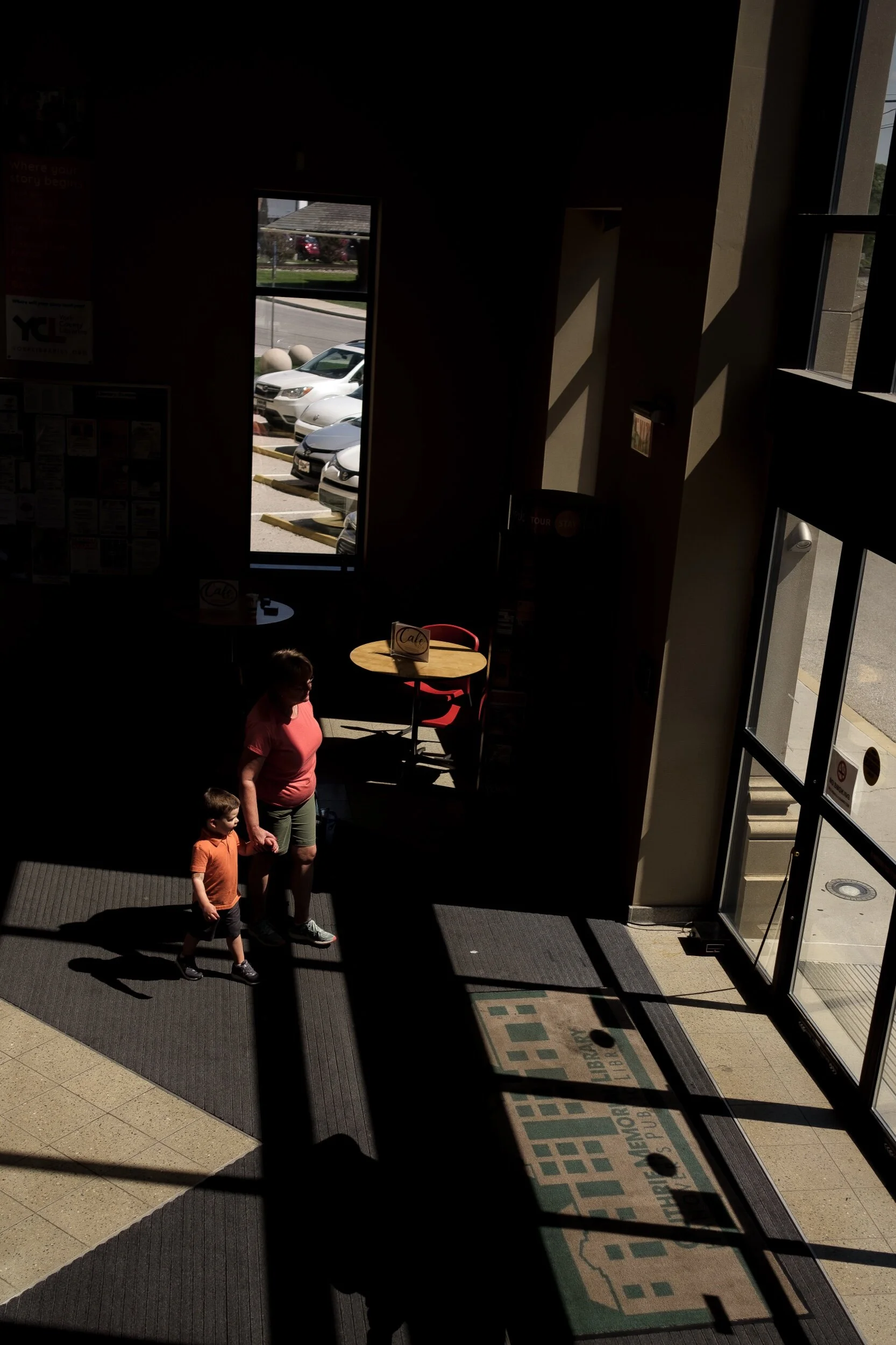 A woman and child walking hand in hand in a dimly lit lobby area with geometric patterns of sunlight and shadows on the floor. A window shows parked cars outside.