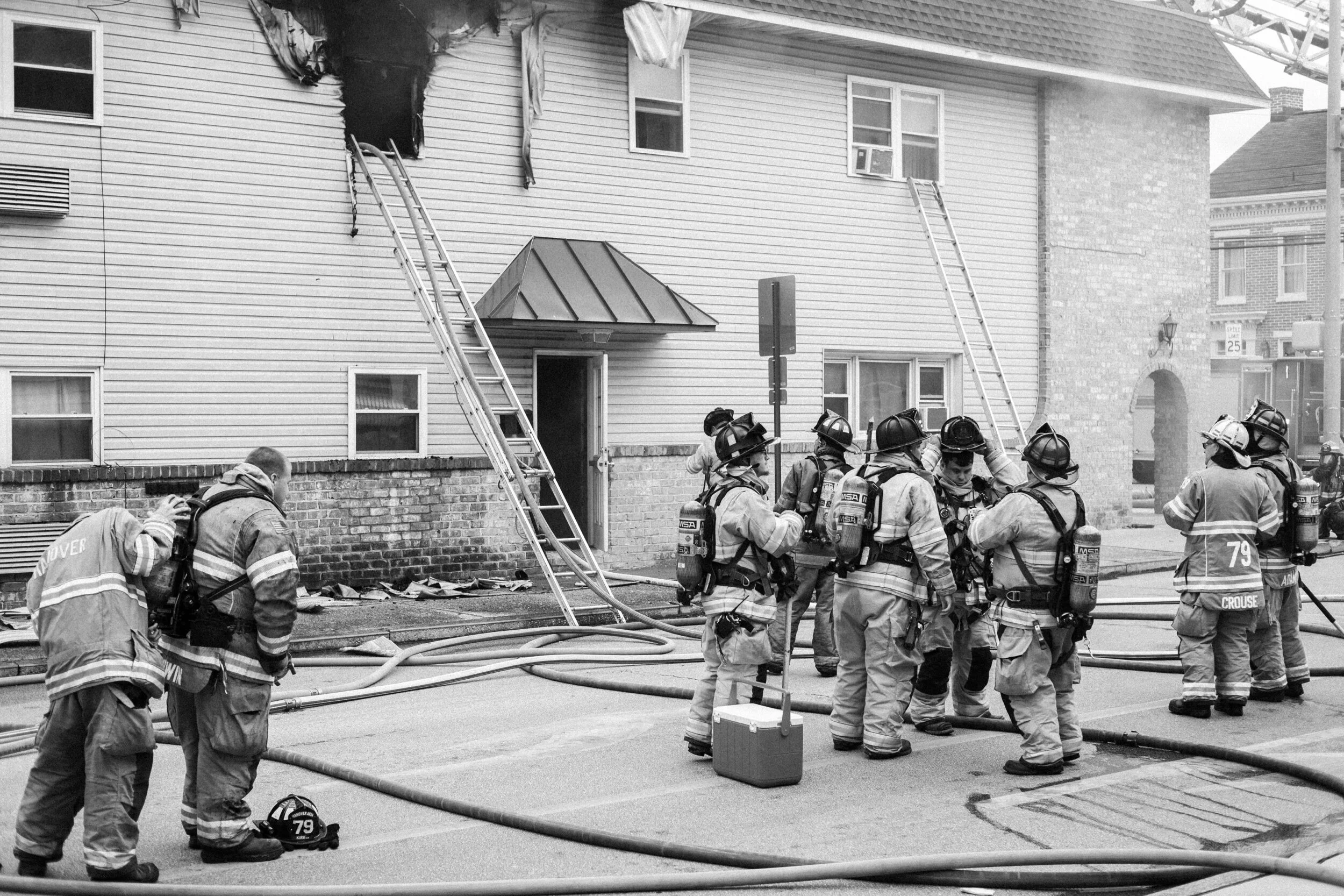 Firefighters wearing protective gear and helmets standing near a building with visible fire damage and ladders, preparing or finishing their work involving firefighting hoses.