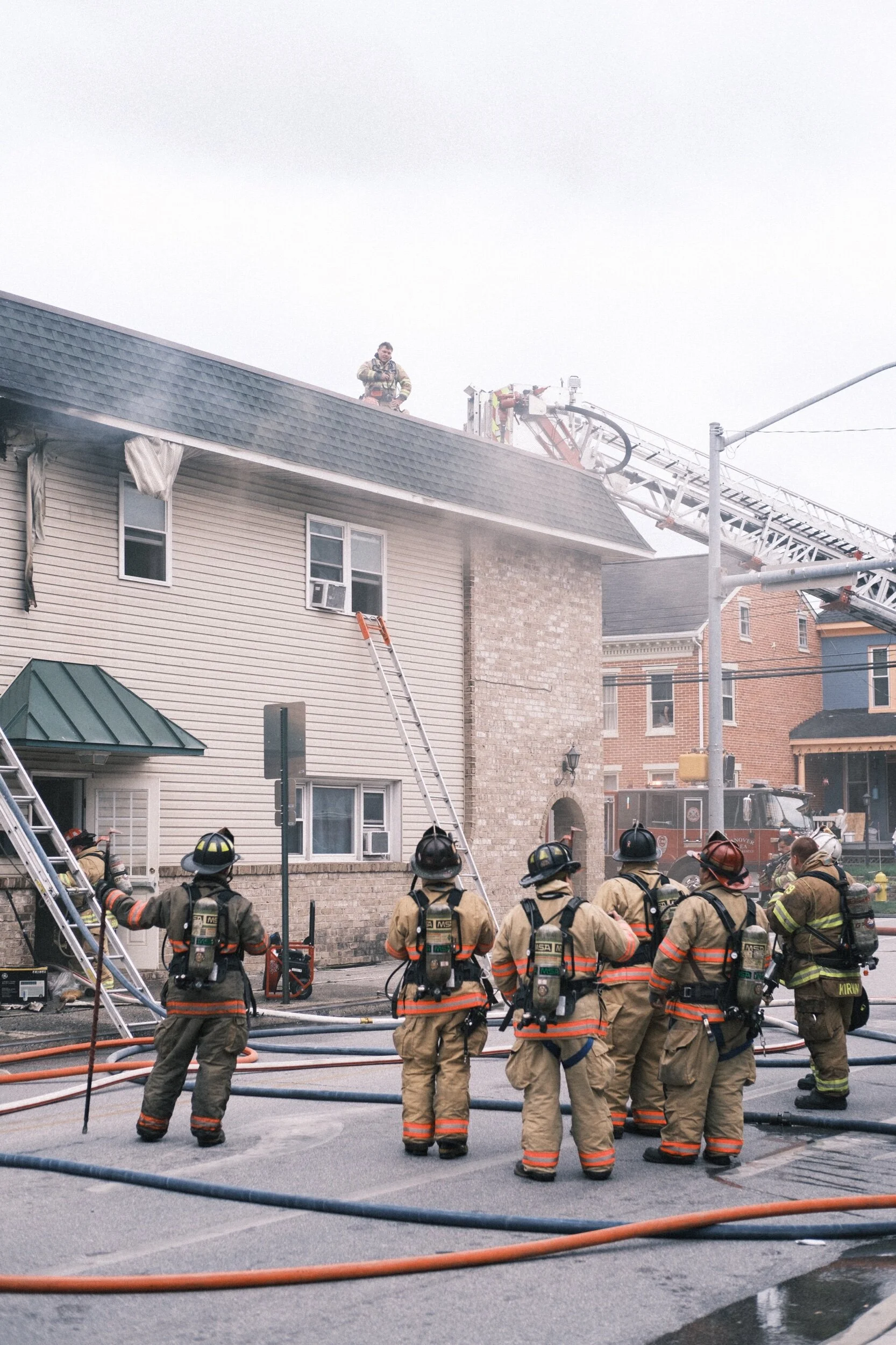 Firefighters in gear respond to a building fire, with ladders against the structure and smoke visible. A fire truck is in the background with more firefighters on the roof.