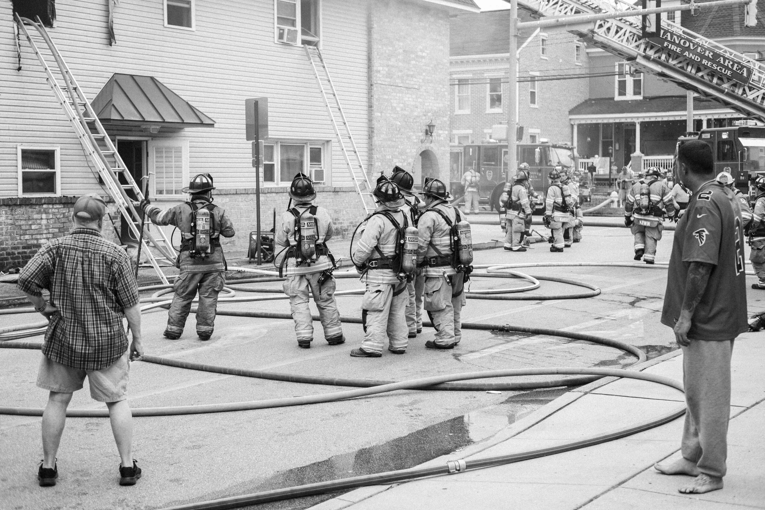 Firefighters in gear handling hoses in front of a building with ladders, observed by onlookers, during a fire emergency in an urban area.