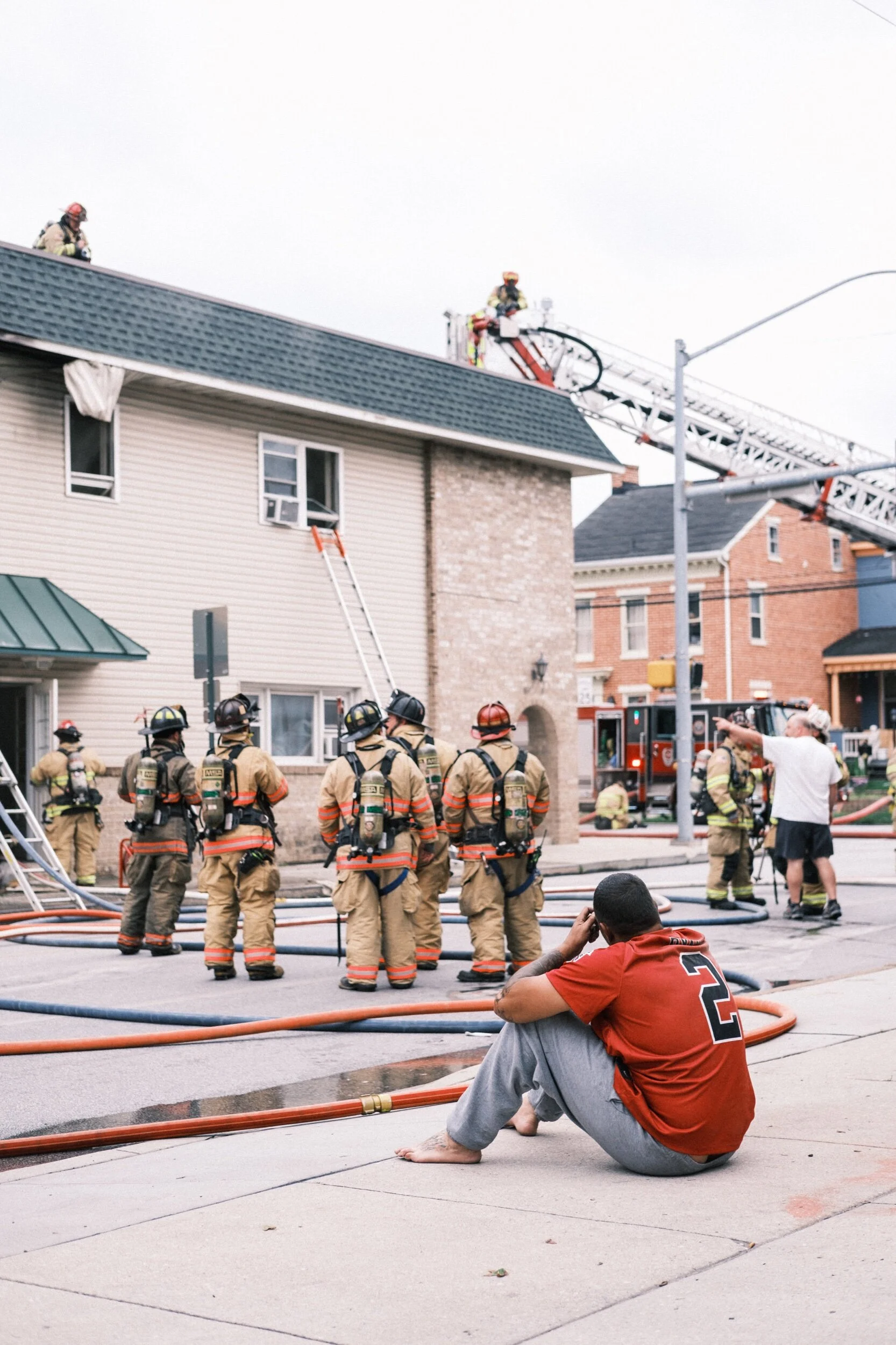 Firefighters in gear responding to a building incident, with ladders and hoses deployed. A person in a red shirt sits on the sidewalk observing the scene.