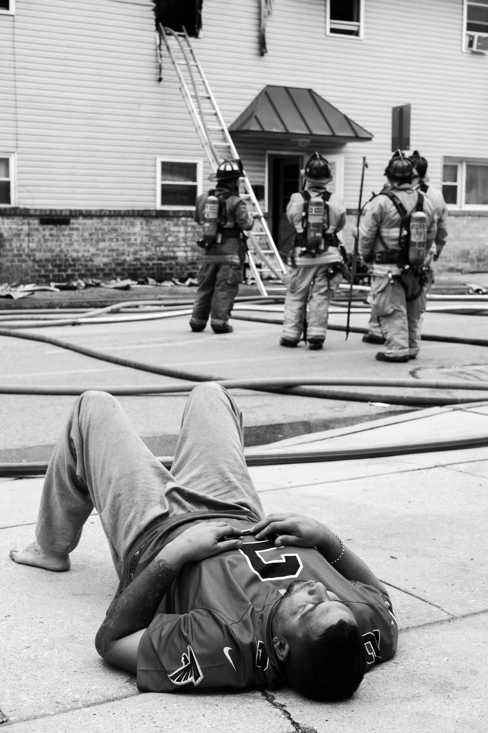 A person lying on the ground in front of firefighters standing by a building with a ladder and hoses, possibly responding to an emergency situation.
