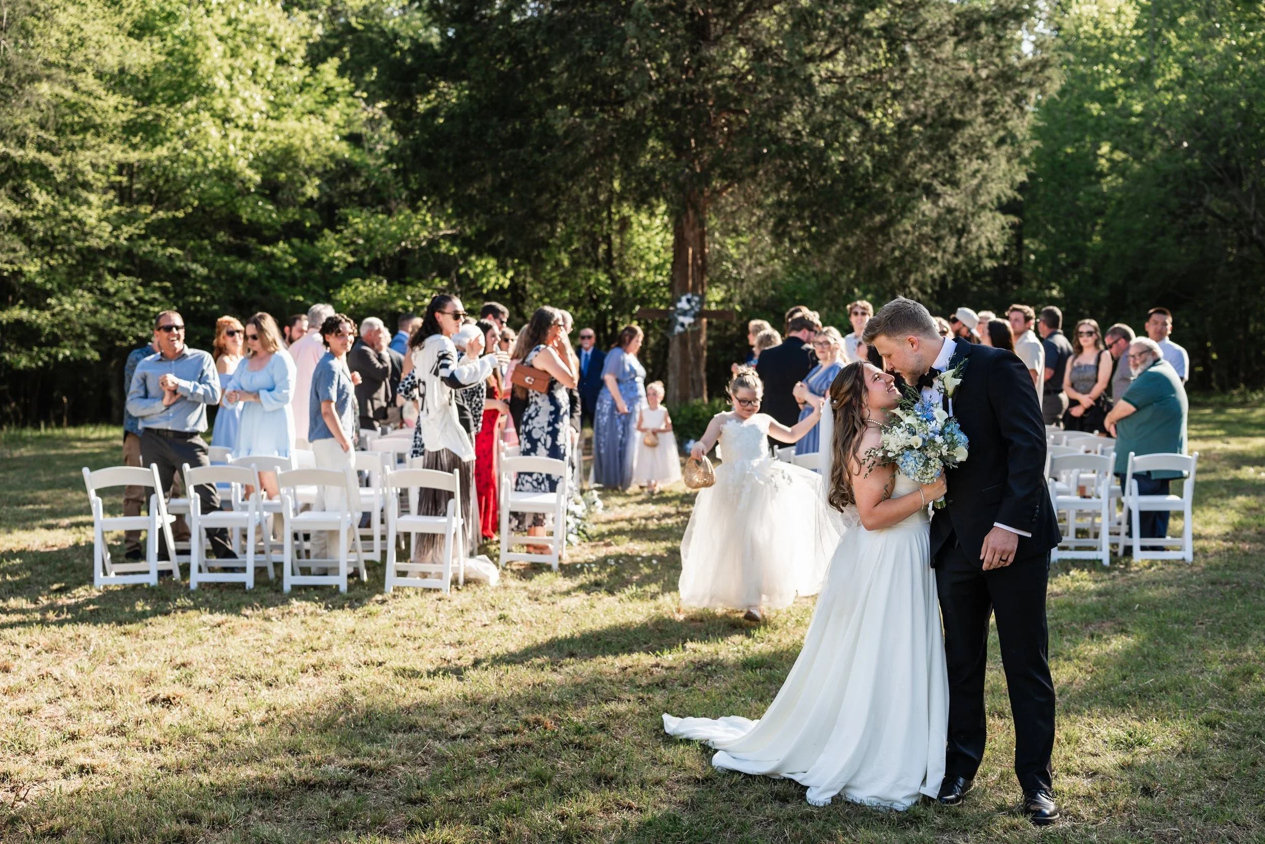 A bride and groom share a moment at an outdoor wedding ceremony with guests seated and standing in the background, surrounded by trees.