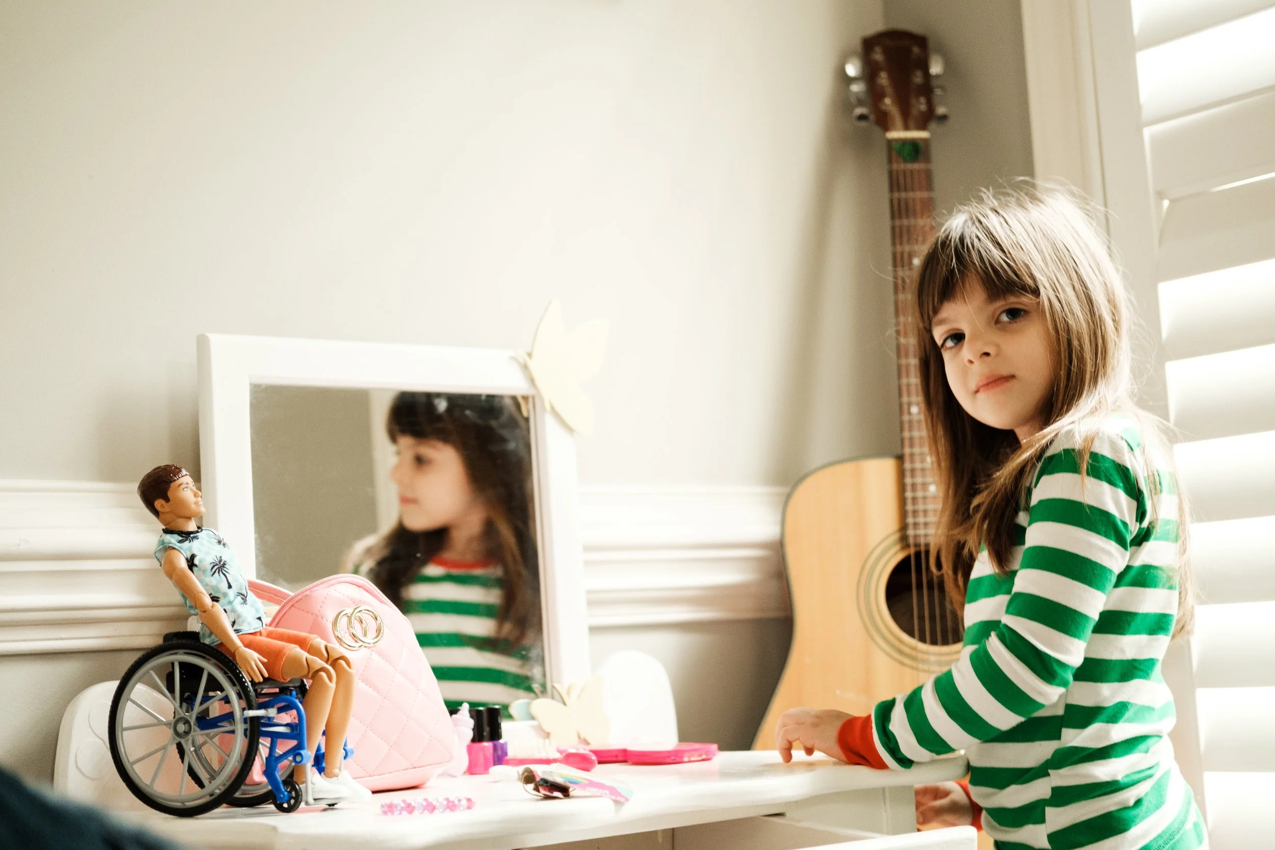 A young girl with long brown hair and a striped green and white shirt is sitting at a white table. On the table, there is a Barbie doll in a wheelchair, a pink handbag, and various small toys and accessories. A mirror reflects her face, and behind her is a beige wall, a window with white blinds, and a guitar hanging on the wall.