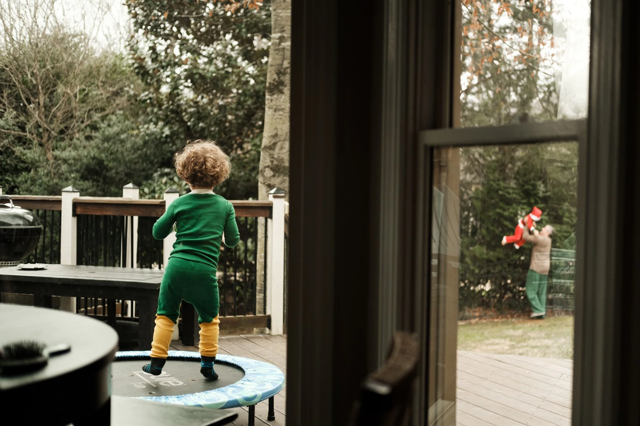 Child with curly hair in green sweater and yellow pants jumping on a small trampoline on a wooden deck outside; viewed from inside through a sliding glass door with Christmas decorations hanging on the outside during family photo shoot in Charlotte.