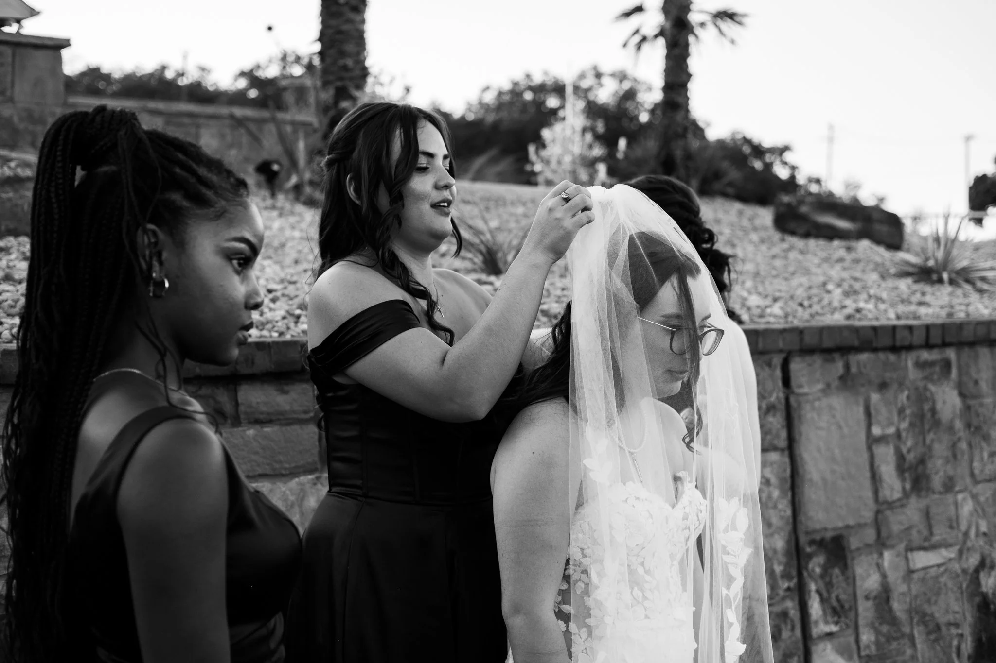 Four women sitting outdoors, with one woman adjusting a veil on the head of another woman wearing glasses; trees and a stone wall in the background.
