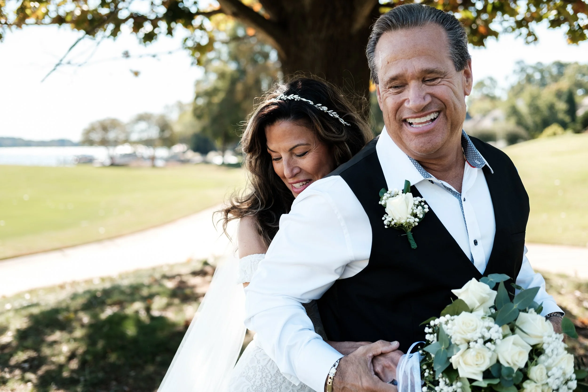 A bride in a white wedding dress and an older man in a suit are smiling and embracing under a tree in a park during a wedding celebration.