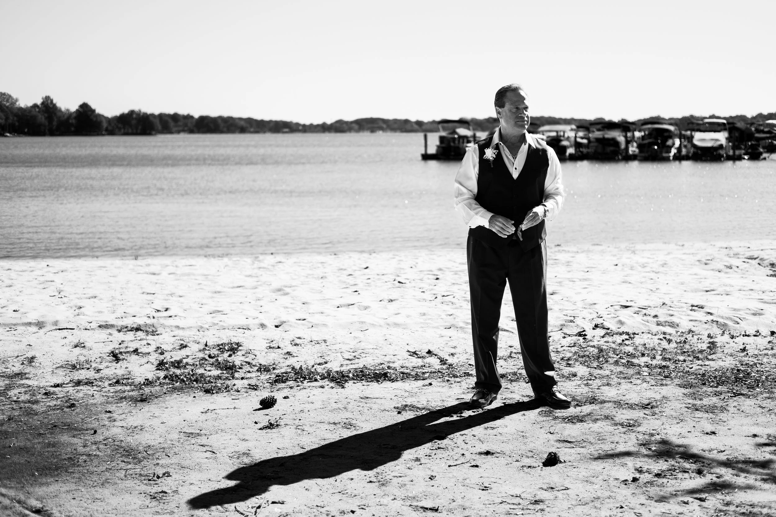 black and white photo of a groom in a tuxedo, waiting in the sand by Lake Norman. He appears to be nervously waiting for the bride to arrive to their wedding ceremony