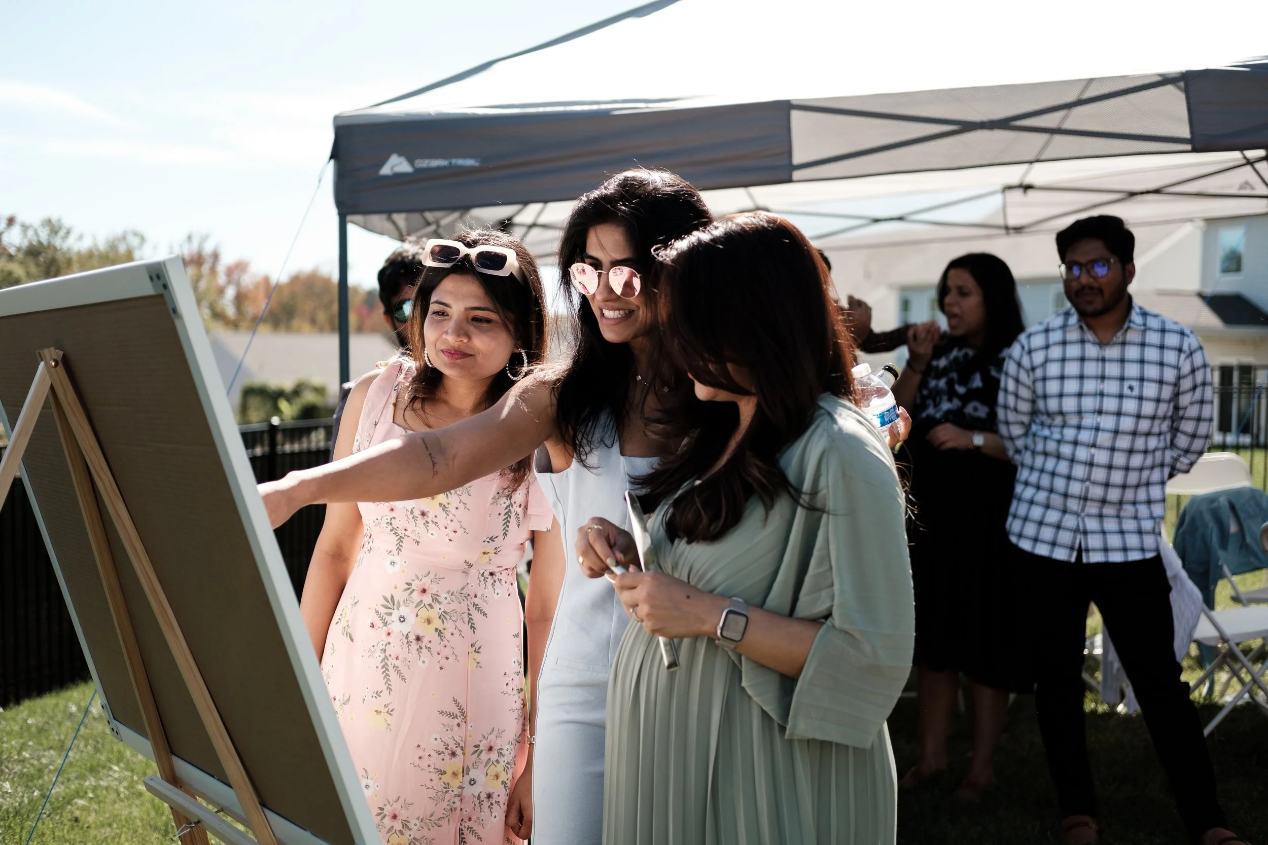 A group of people at an outdoor event by Charlotte photographer Brandon Pickett under a canopy. Three women are focused on a board or canvas, one is pointing, while others stand in the background. The setting is sunny, with grass and trees visible.