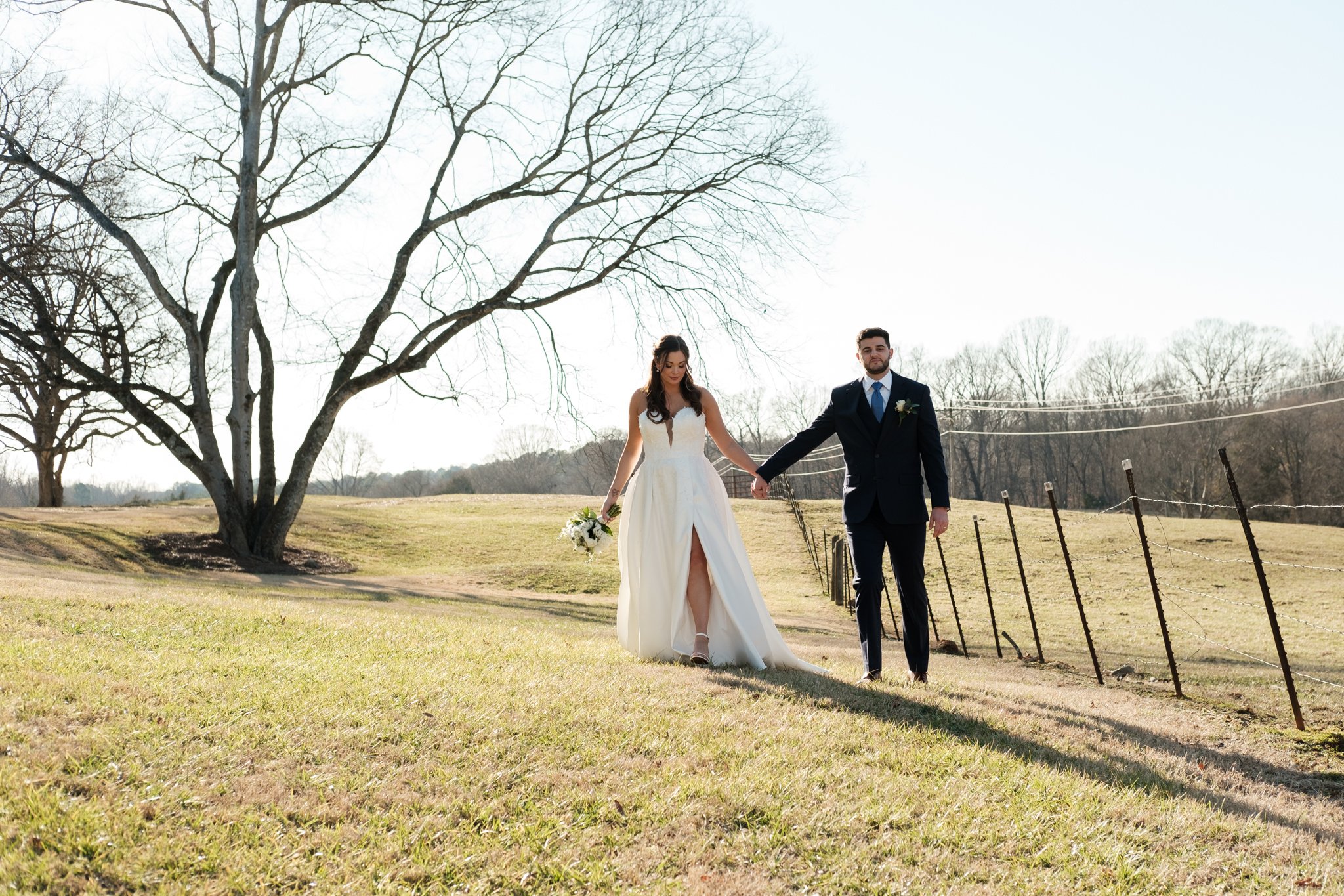 A bride in a white wedding gown and a groom in a dark suit walk hand in hand outdoors in a grassy area at Founders Dairy Barn.