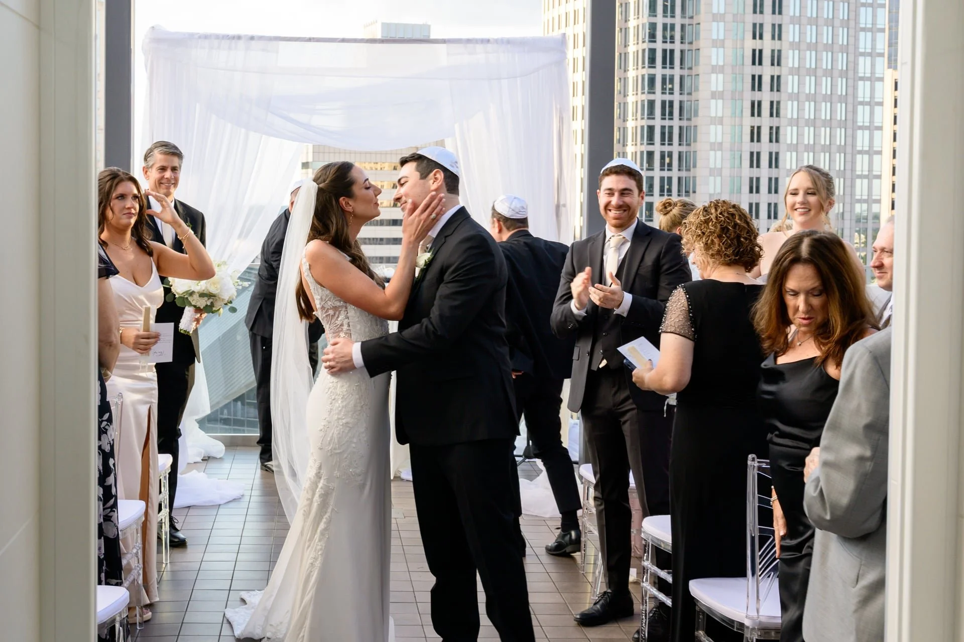 A wedding ceremony on a rooftop, with the bride and groom embracing and surrounded by family and friends, city skyline in the background.
