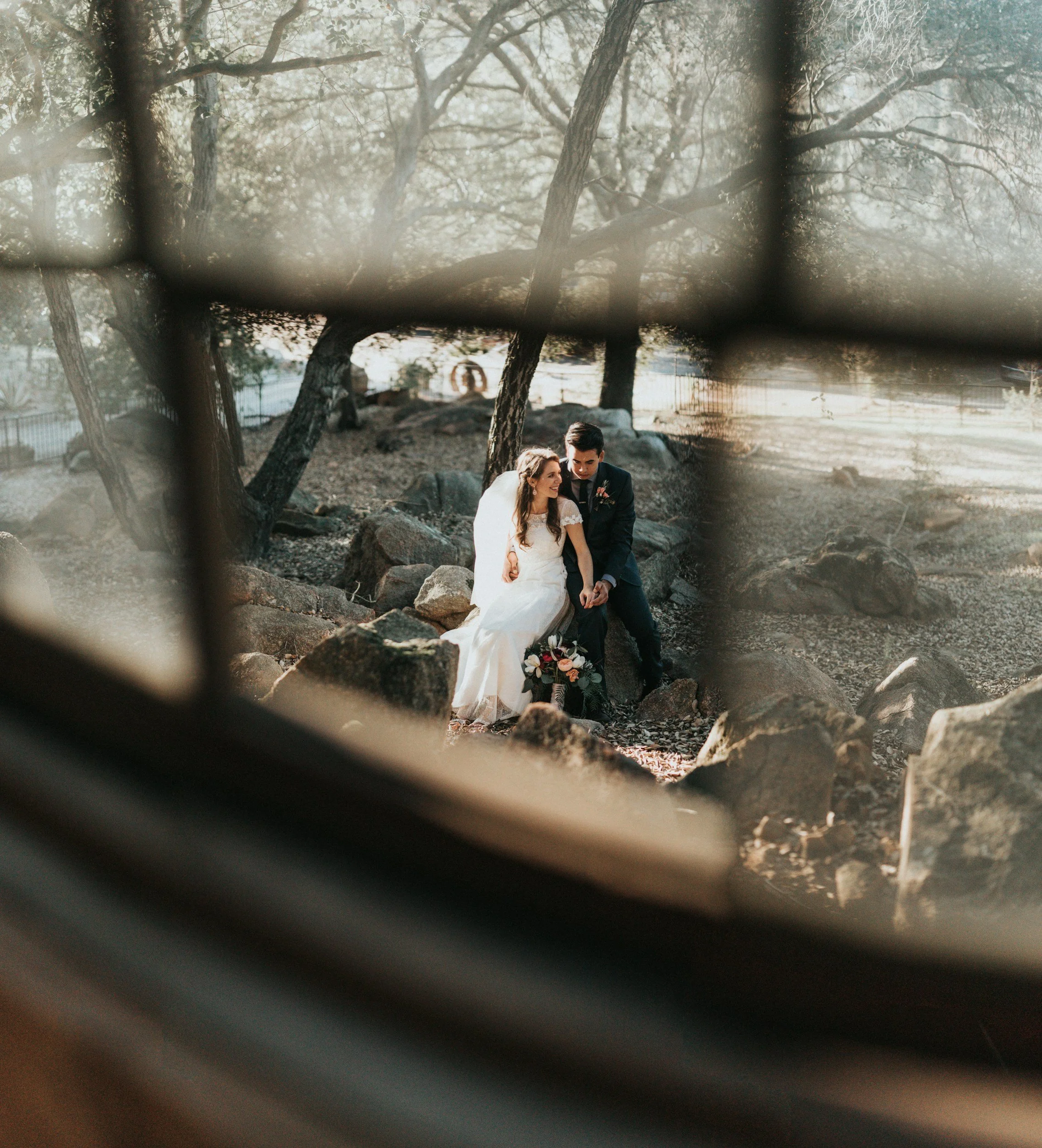 bride and groom sitting on a rock smiling. Photo shot through an old window in Charlotte, NC.