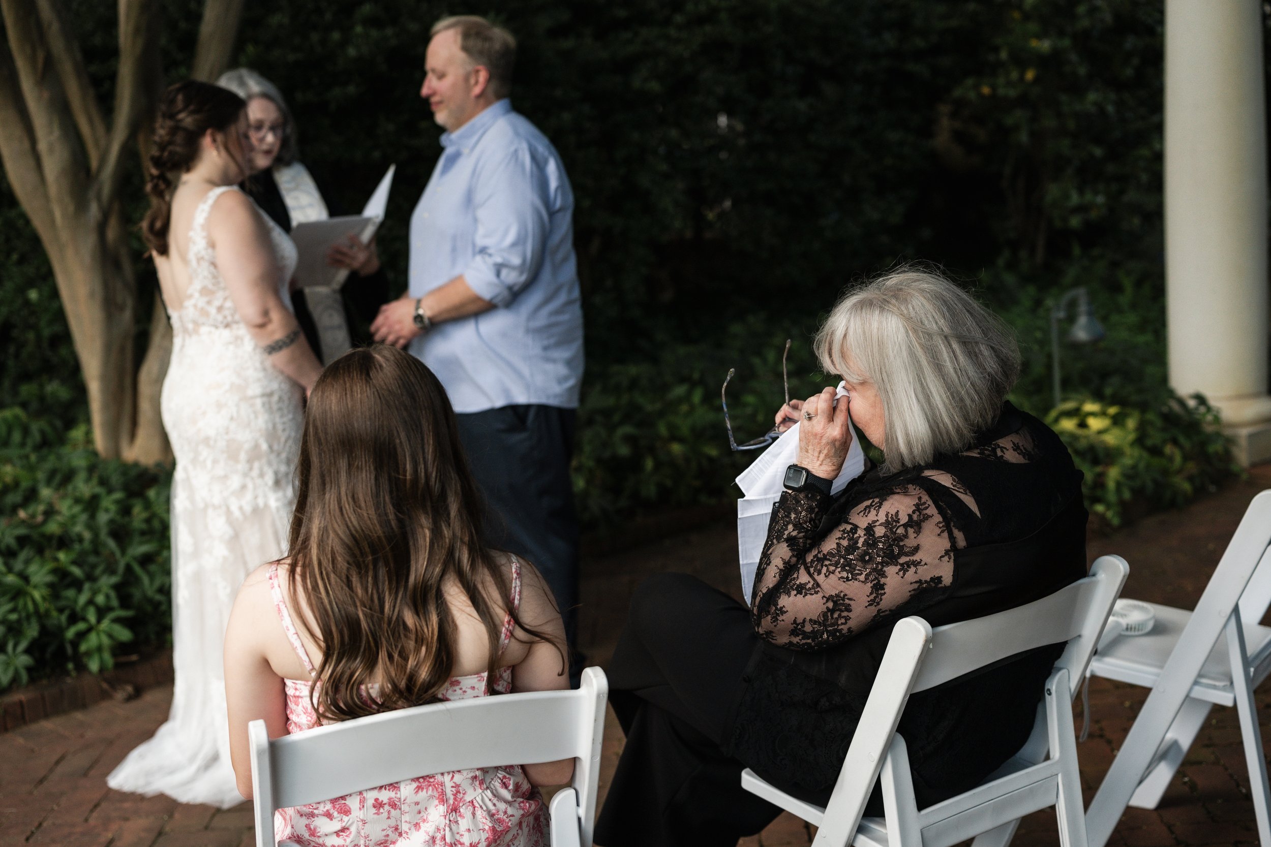 A wedding ceremony at Daniel Stowe, with a bride in a lace white dress and a groom in a light blue shirt, reading vows. An officiant stands behind them, and a woman with gray hair and black lace attire is seated, wiping tears or eyes with a tissue.