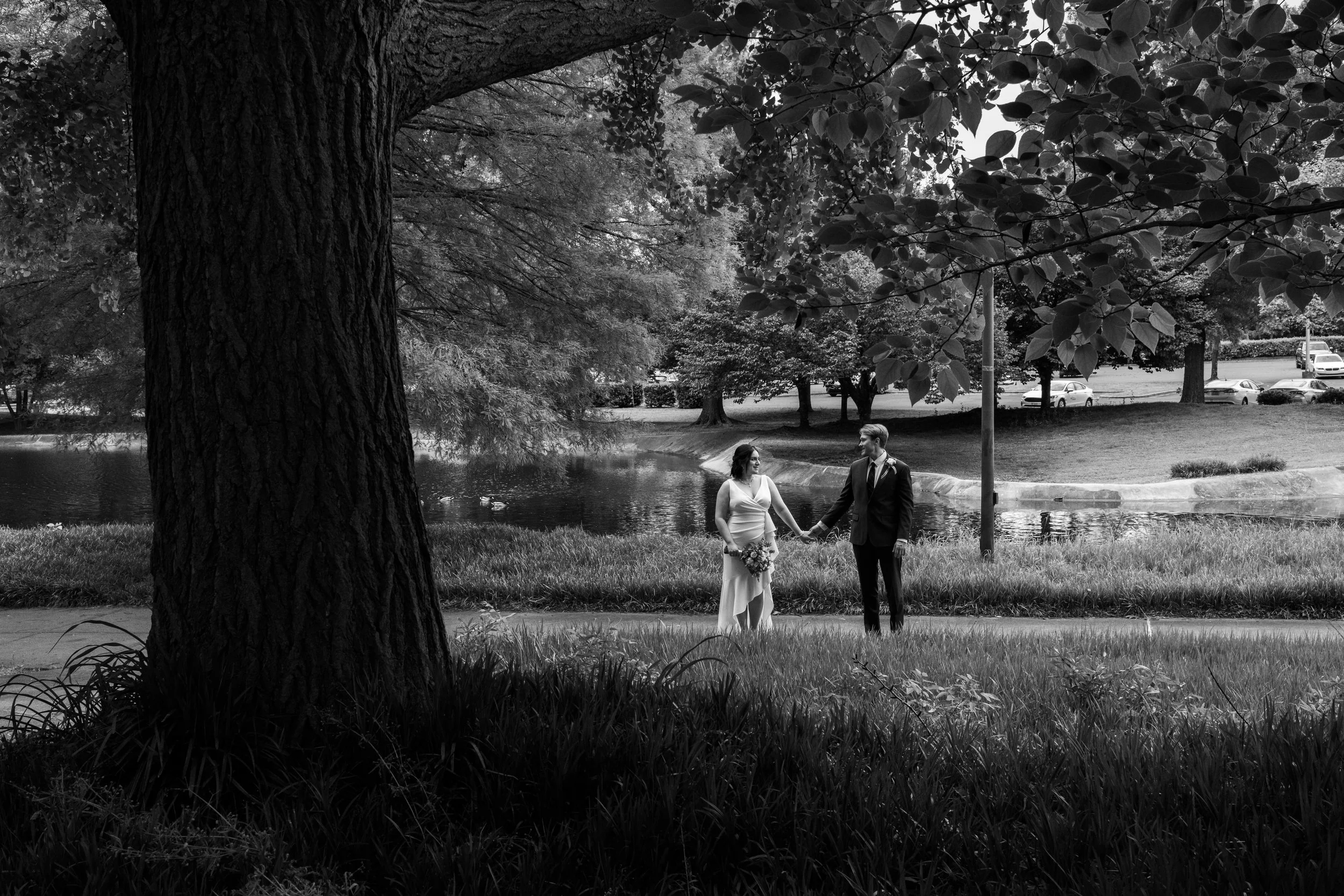 A black and white photo of a couple holding hands near a pond in a park, with large trees and cars in the background.