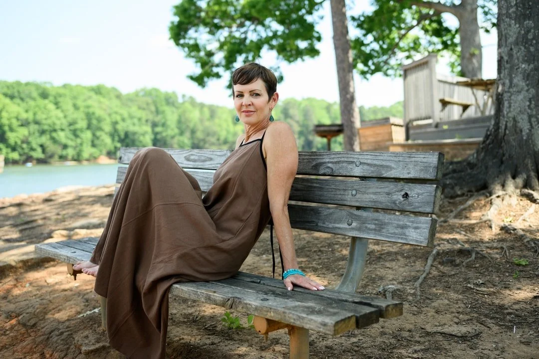 Woman sitting on a wooden park bench near the water with trees in the background during a portrait photography session in Charlotte, NC.