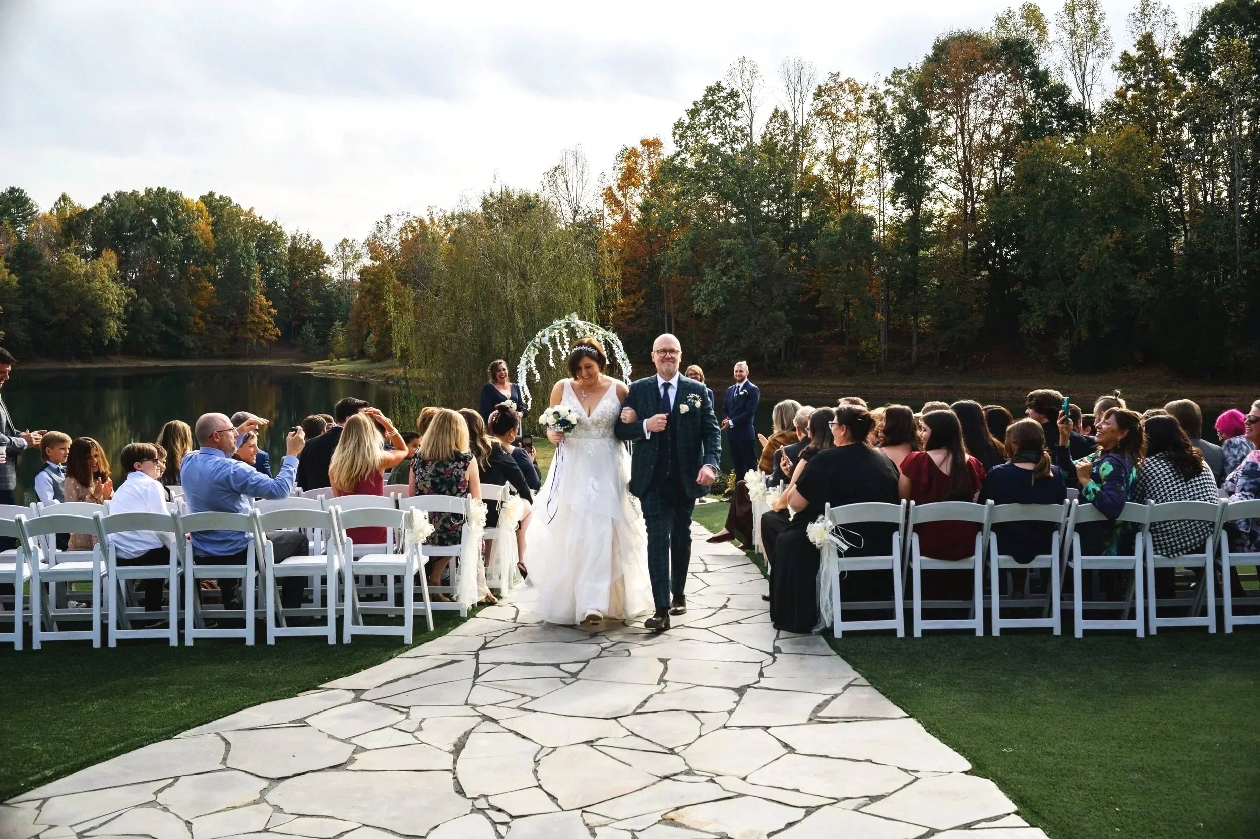 A wedding ceremony at Splendor Pond Flower Farm, featuring a bride and a groom walking down the aisle, surrounded by seated guests, in a natural setting with an arch decorated with flowers in the background.