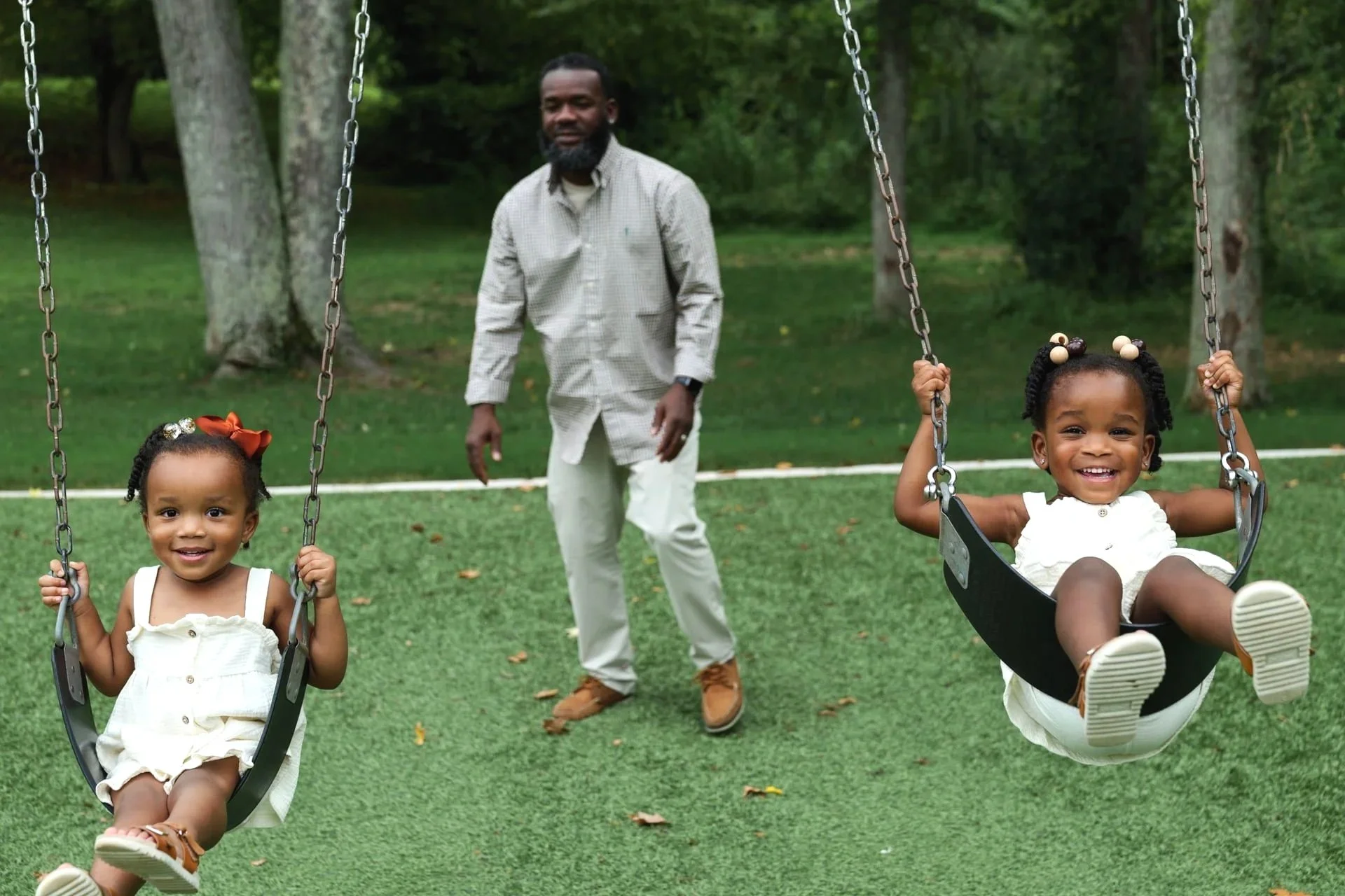 Two young girls playing on swings in a park with a man standing behind them, surrounded by green grass and trees.