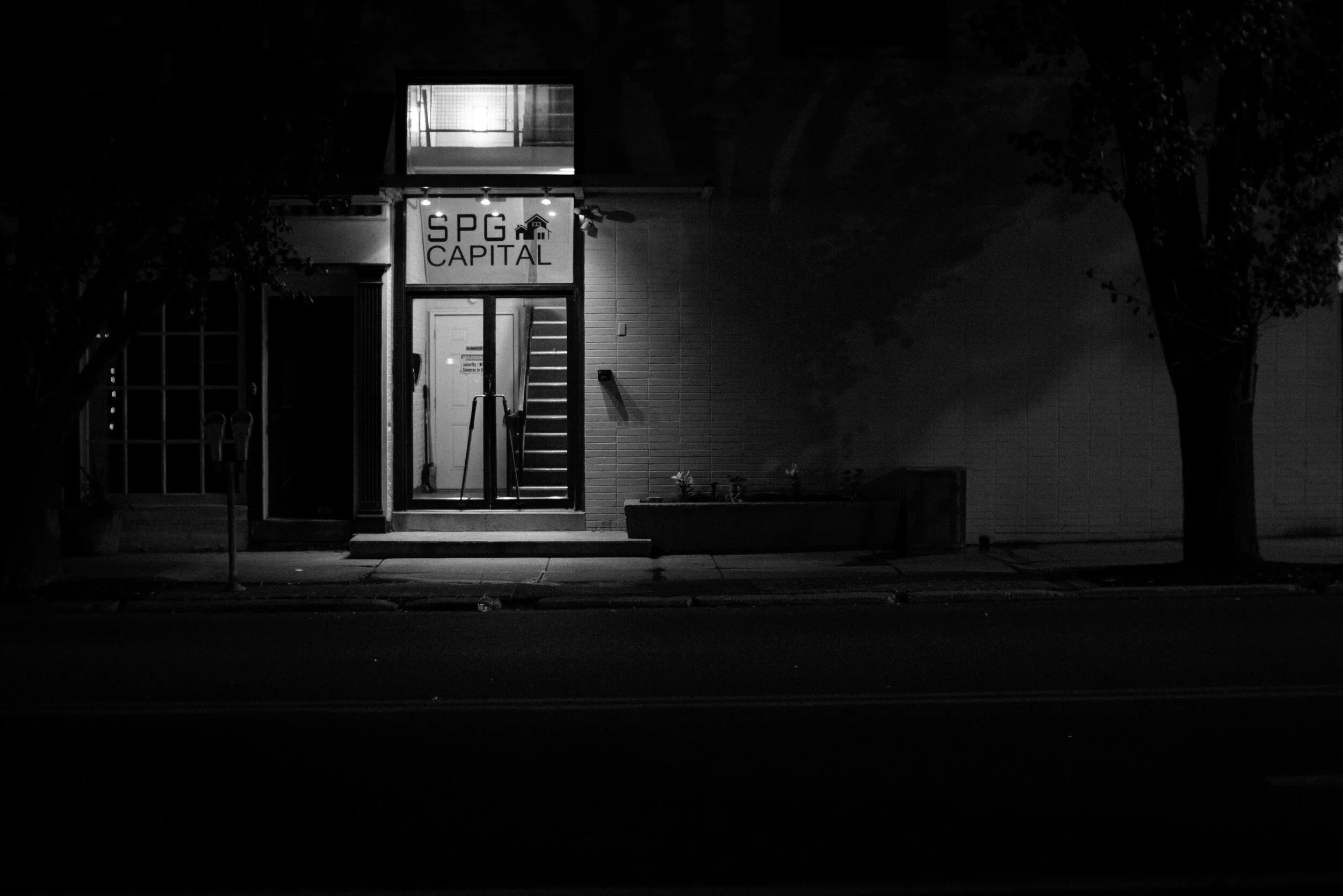 Black and white photo of an office entrance with "SPG Capital" sign above the door, night view, dim lighting.
