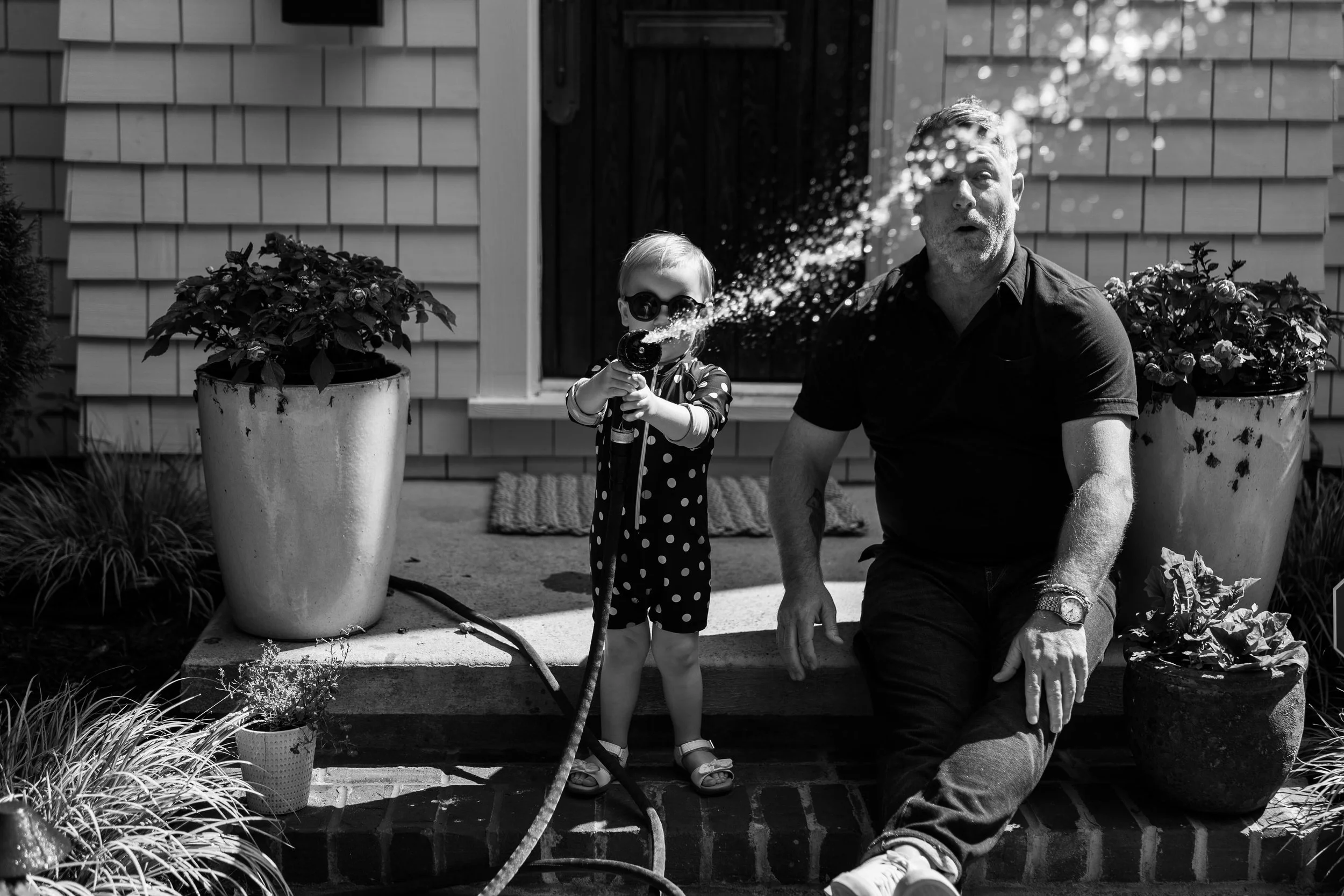 A young child wearing sunglasses and a polka dot dress holds a garden hose, spraying water which creates a mist in the air, while an older man with a beard and short hair sits on a brick ledge beside her. They are outdoors in front of a house with shingle siding, plants, and large pots with flowers.