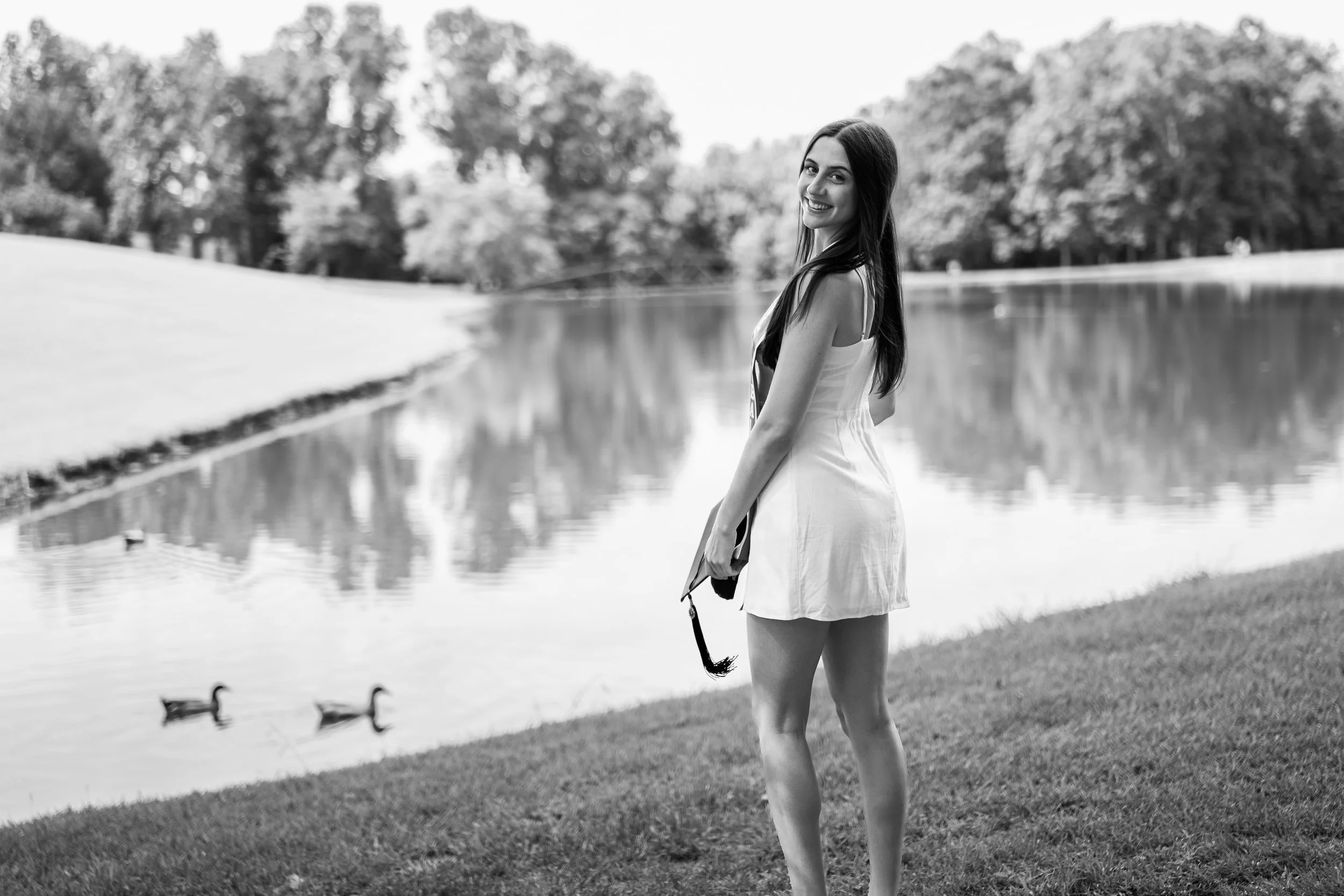 A woman in a white dress standing by a lake with ducks swimming, surrounded by trees, smiling and looking back at the camera.
