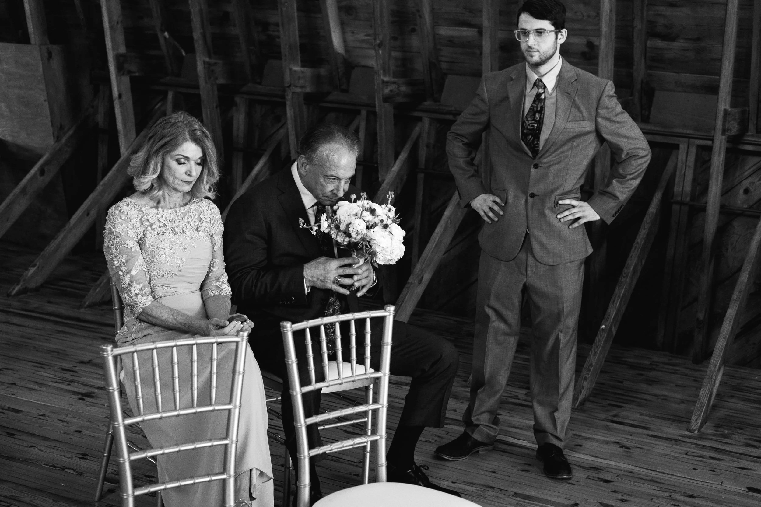 Three people are sitting and standing in a rustic wooden space, with two women and one man. The woman on the left has light hair and wears an ornate dress, looking down with her hands clasped. The man in the middle, holding a bouquet of flowers, has gray hair and a dark suit. The man on the right has dark hair, glasses, and a light gray suit with a patterned tie, standing with hands on hips.
