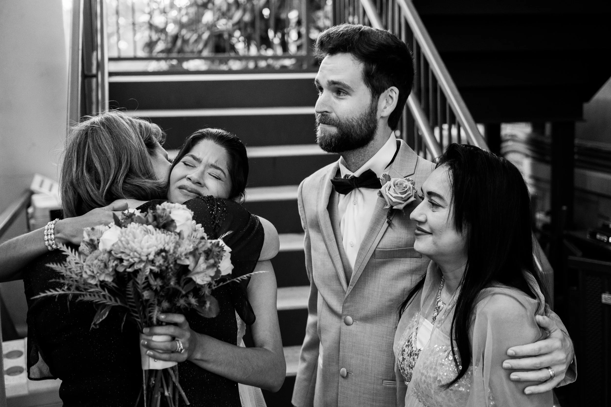 A black and white photo of a man in a suit with a boutonniere, embracing and consoling a woman holding a bouquet, with three other women nearby, on a staircase with a railing and some plants in the background.