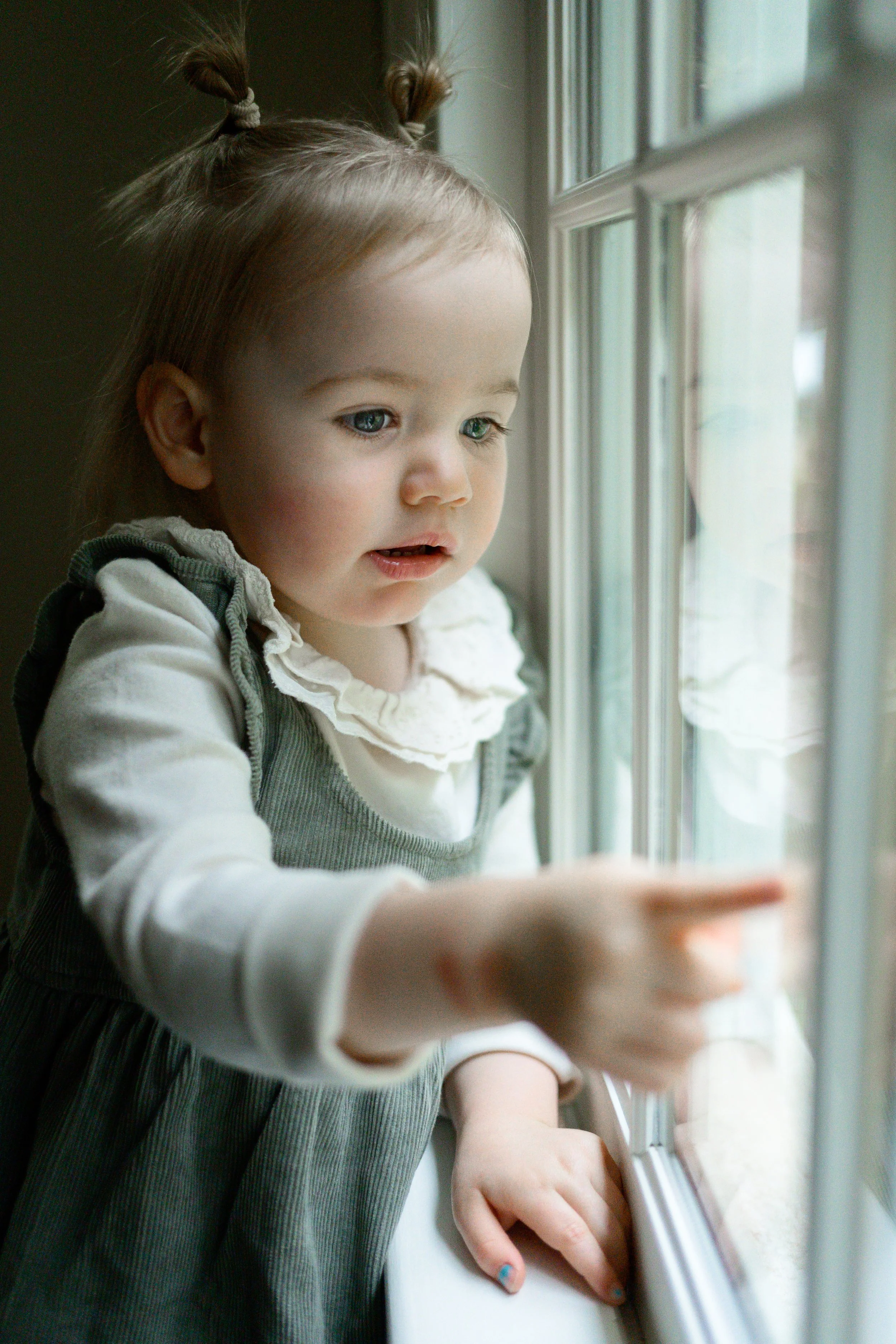 A young girl with light hair and blue eyes, dressed in a grey dress with a white ruffled collar, looking out the window and pointing at something outside.
