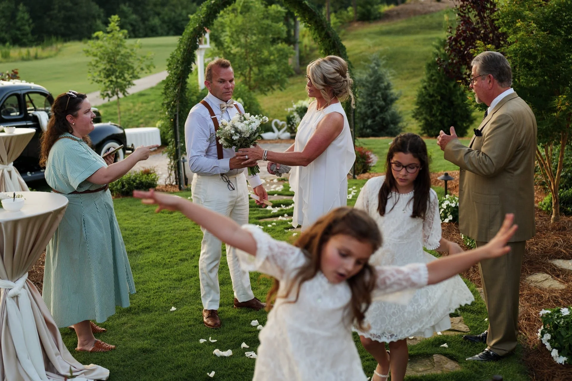 A wedding ceremony outdoors with a bride and groom exchanging flowers, surrounded by family and friends, on a green lawn with trees and vintage cars in the background.