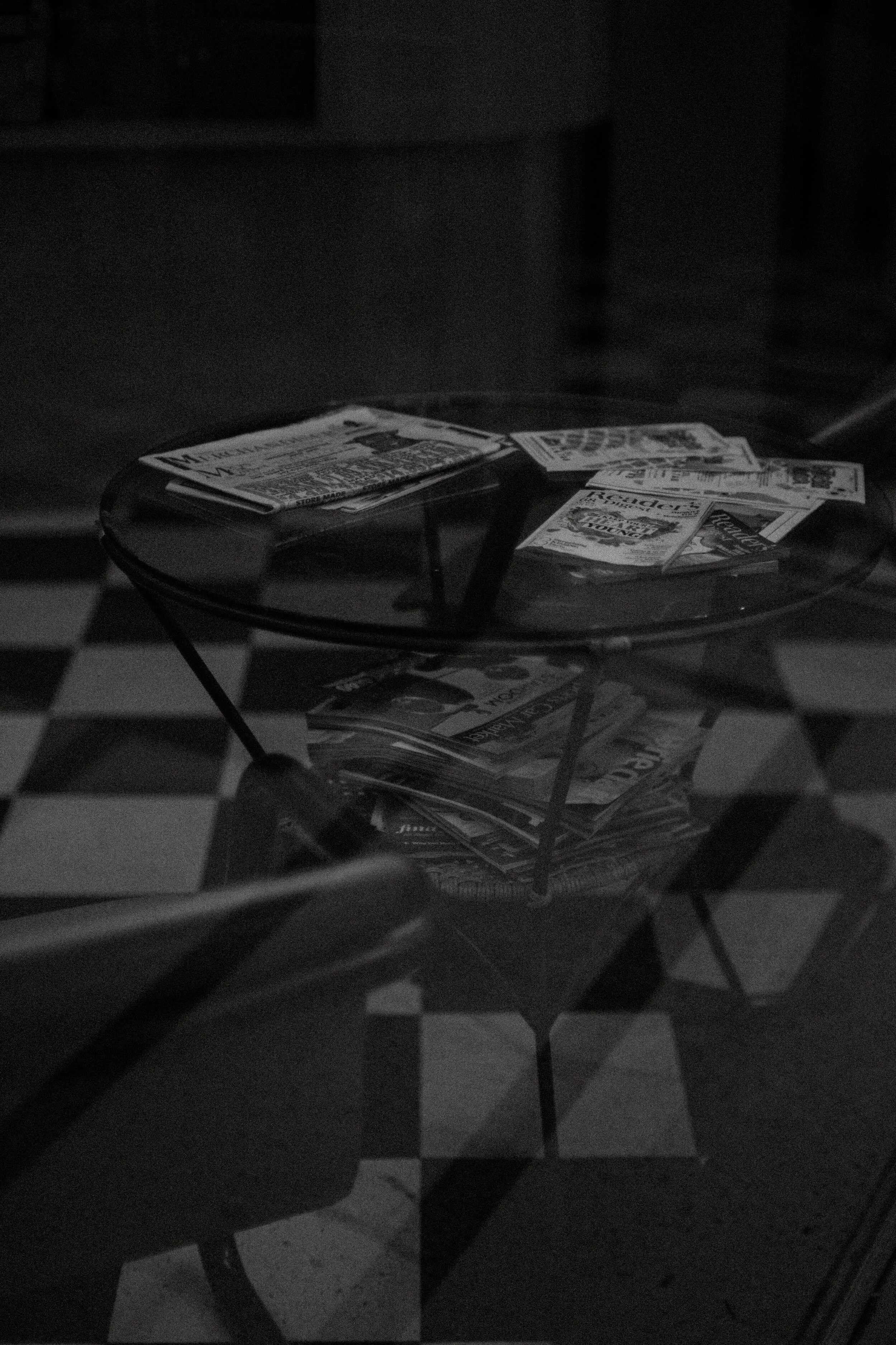 Black and white photo showing a glass-top coffee table with magazines or papers on it, in a room with checkered flooring.
