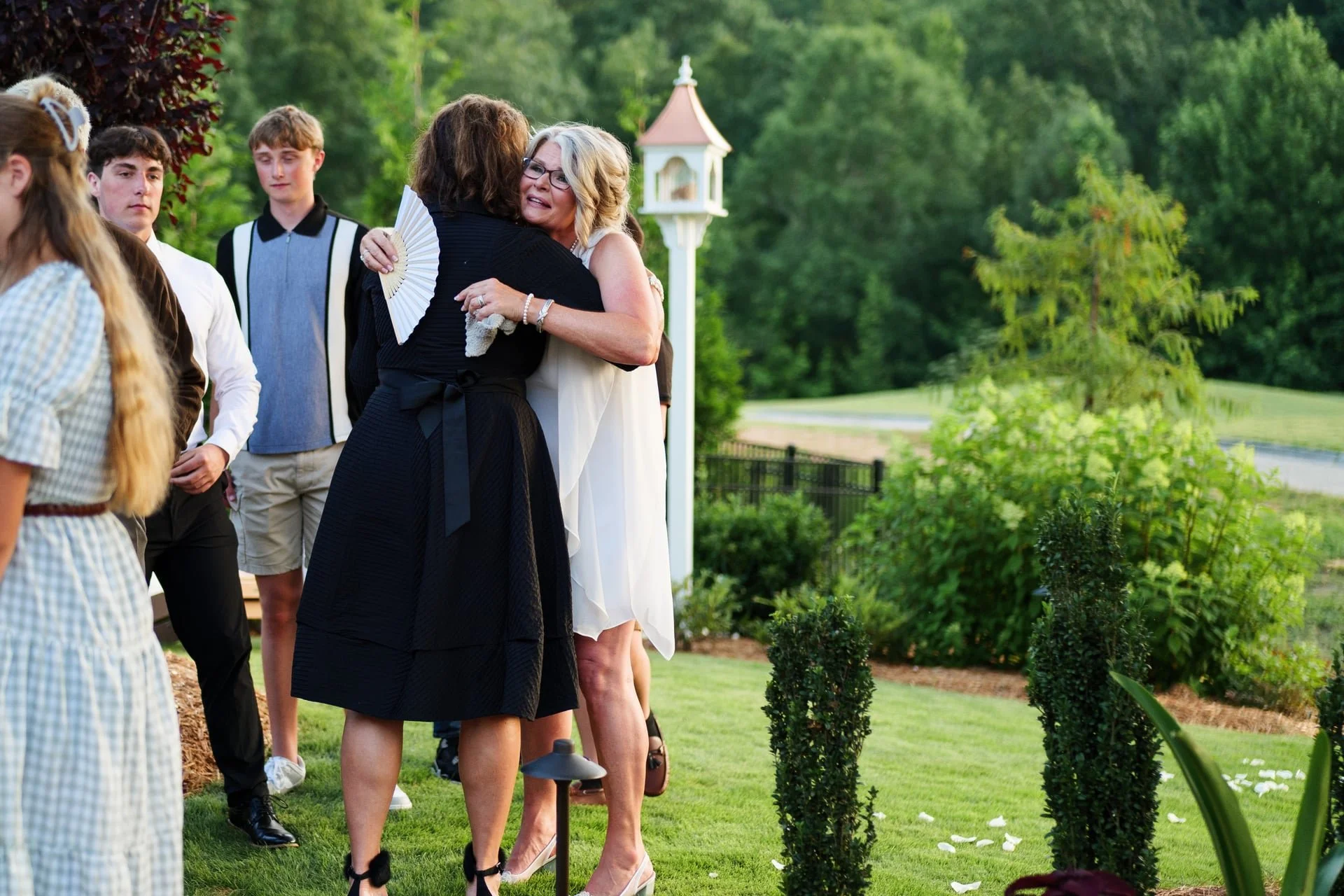 Two women hugging at an outdoor gathering, surrounded by a few young people, with trees and a small building in the background.