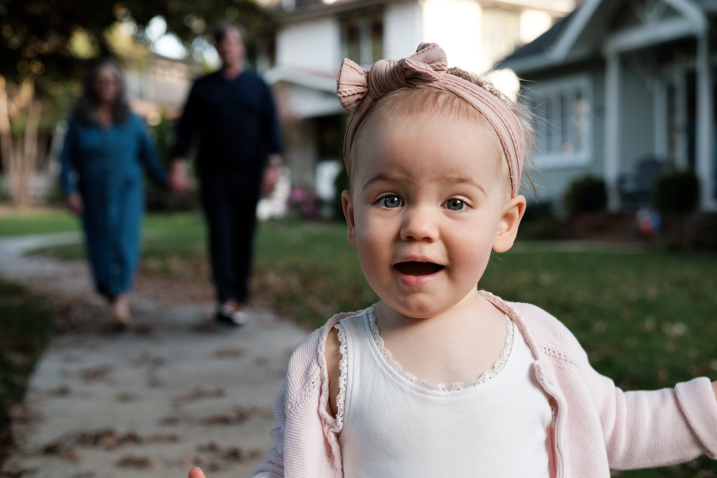 Close-up of a young girl with blue eyes, blonde hair, wearing a pink bow headband and white and pink clothing, outdoors with two blurred adults holding hands in the background.