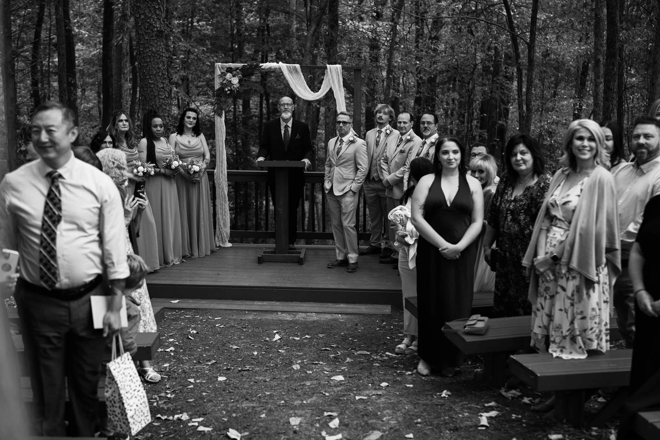 Wedding ceremony in a forest with a group of guests standing on a wooden platform. The officiant is at the podium, and bridesmaids and groomsmen are lined beside him. Guests are seated and standing, some holding bouquets, with trees in the background