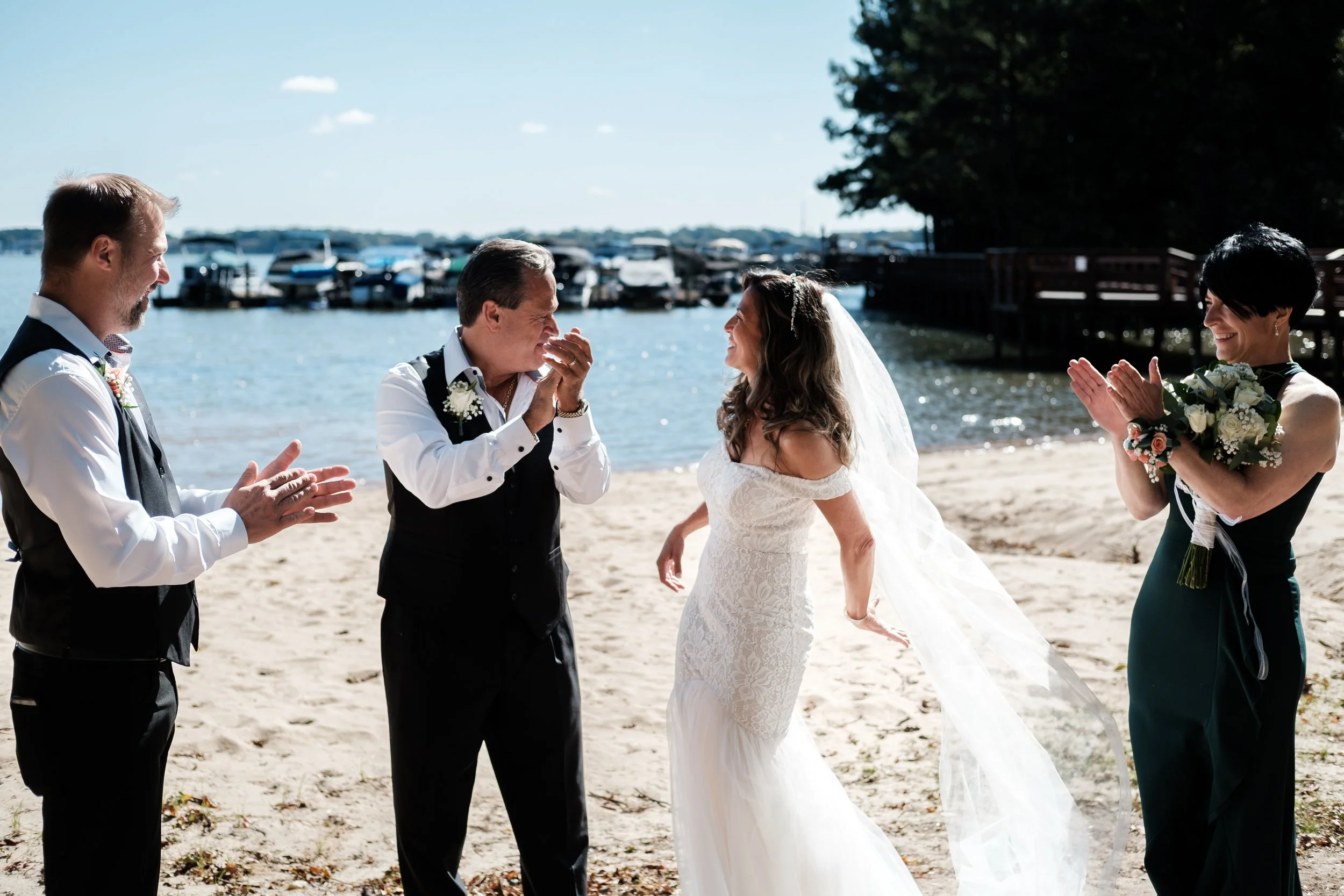 A wedding ceremony taking place on a beach with five people, including two men and two women, and a bride in a white wedding dress and veil. The bride is smiling as the two men, likely her father and another guest, are emotional and smiling. The woma