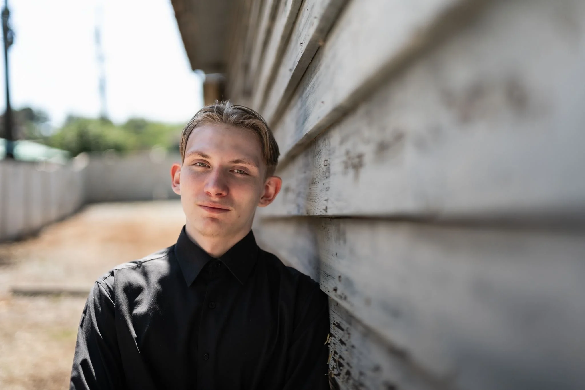 A young man with blond hair wearing a black shirt, leaning against a weathered wooden wall outdoors on a sunny day.