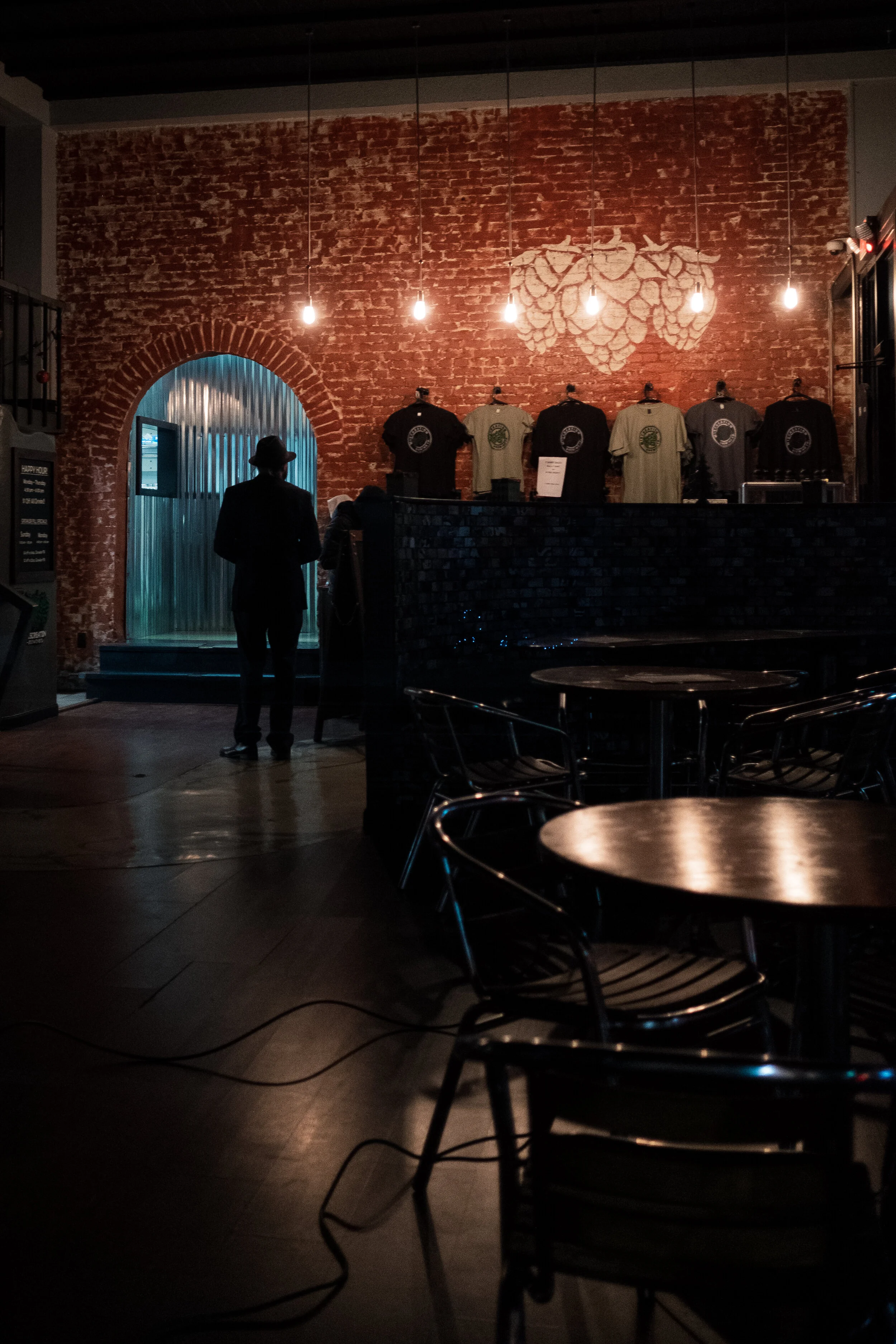 Interior of a dimly lit restaurant with exposed red brick walls, hanging lightbulbs, and a decorative hops design. Tables with metal chairs are in the foreground, and a man in a hat stands near the entrance. T-shirts are displayed on a wall-mounted r