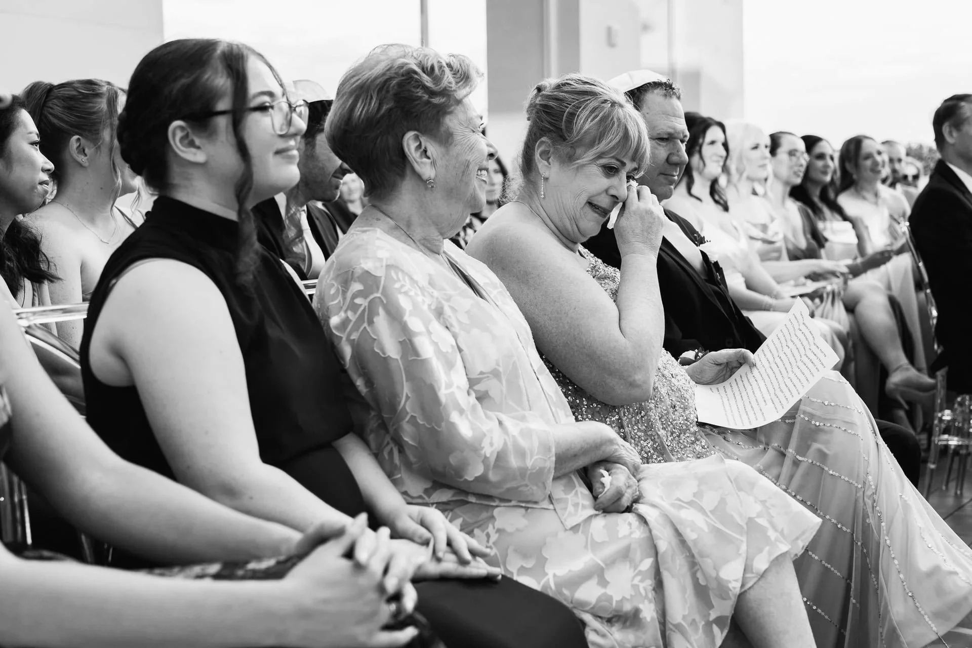 A group of diverse people sitting in an audience, with some smiling and one woman visibly emotional, attending a wedding at Nuvole Rooftop in Charlotte