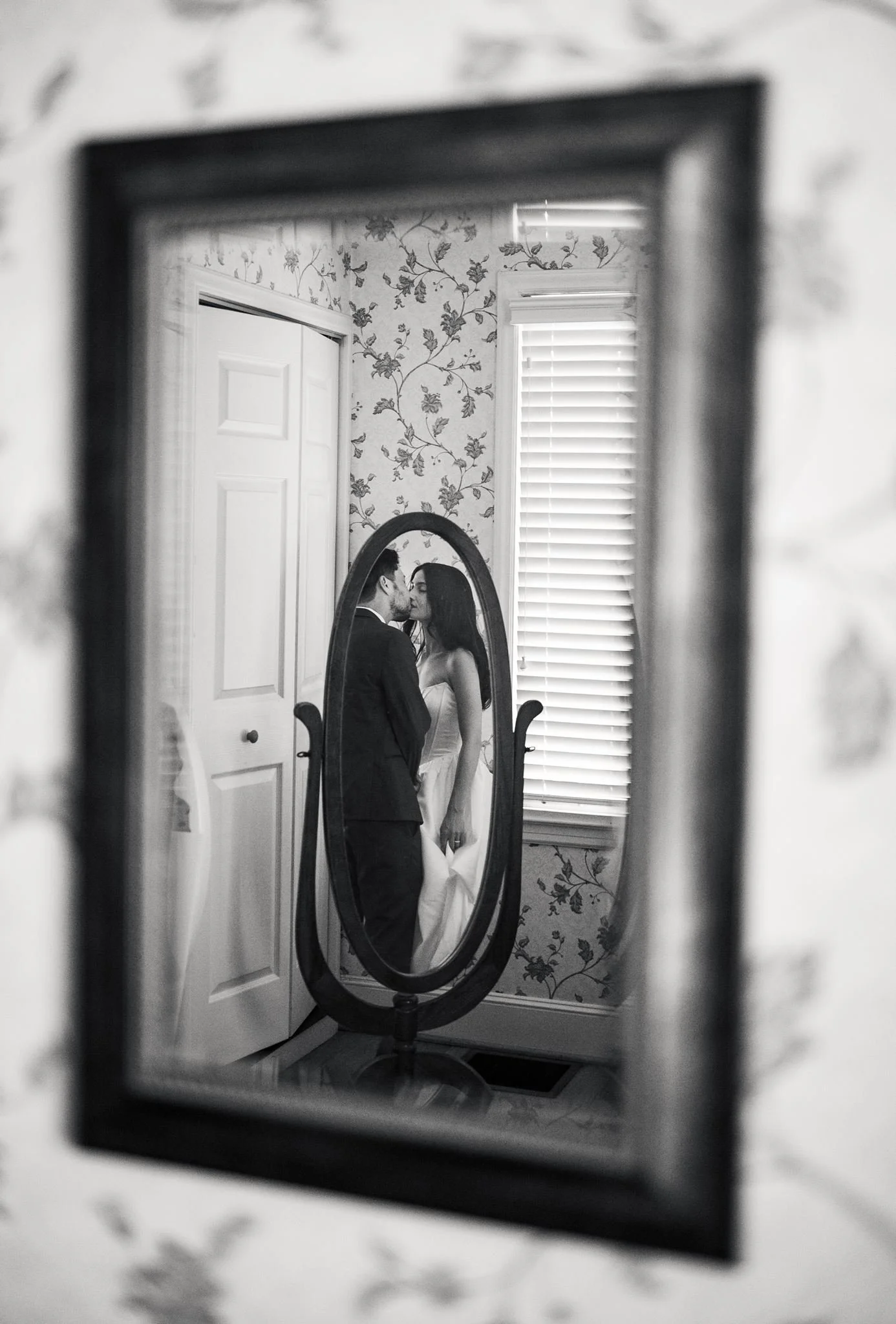 A couple dressed in wedding attire sharing a kiss, seen through the reflection in a mirror in a room at Reid House in Mathews, NC, with floral wallpaper and a window with blinds.