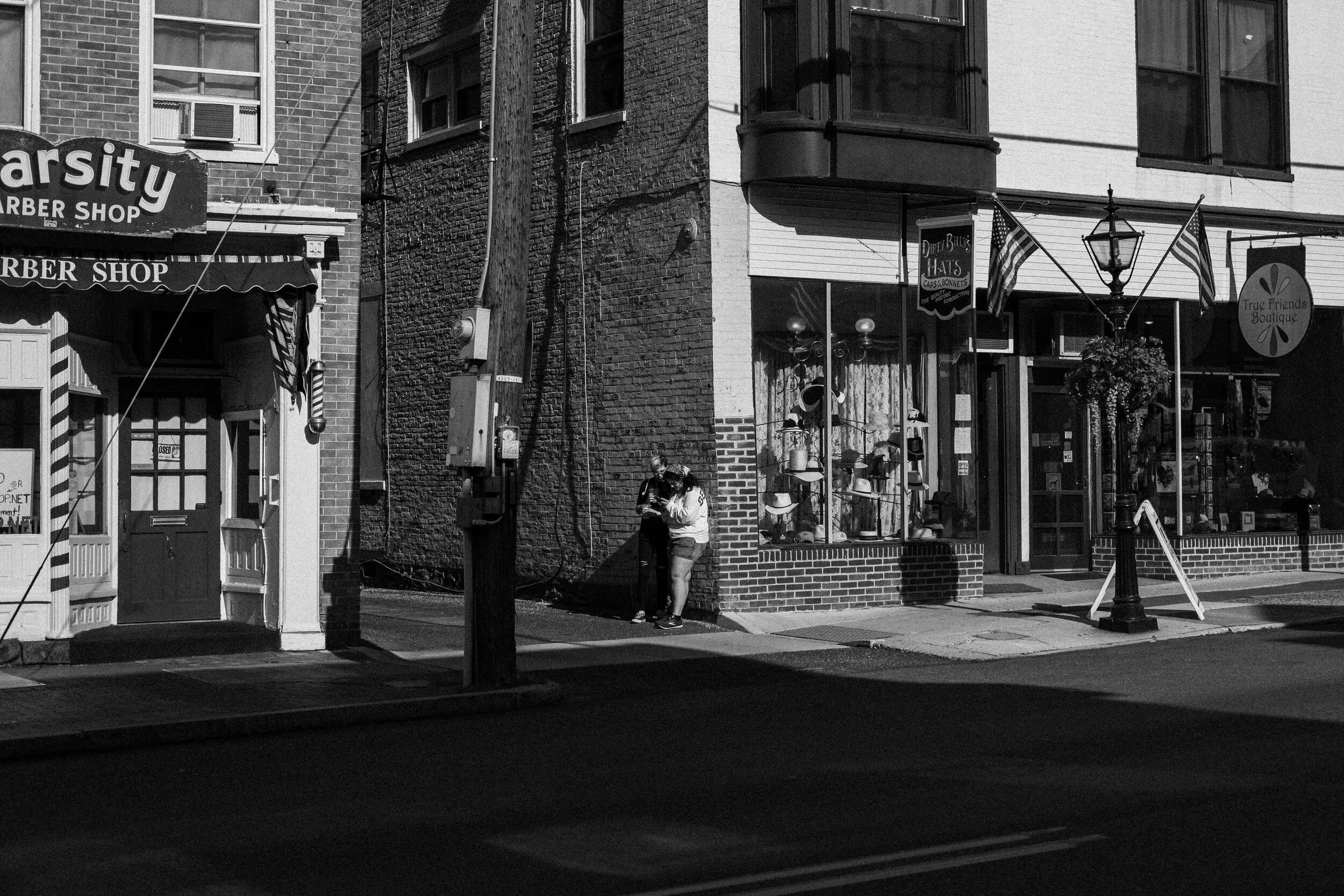Black and white photo of a street corner with a barber shop and boutique. Two people stand on the sidewalk near hats displayed in a shop window. American flags hang outside the shops.