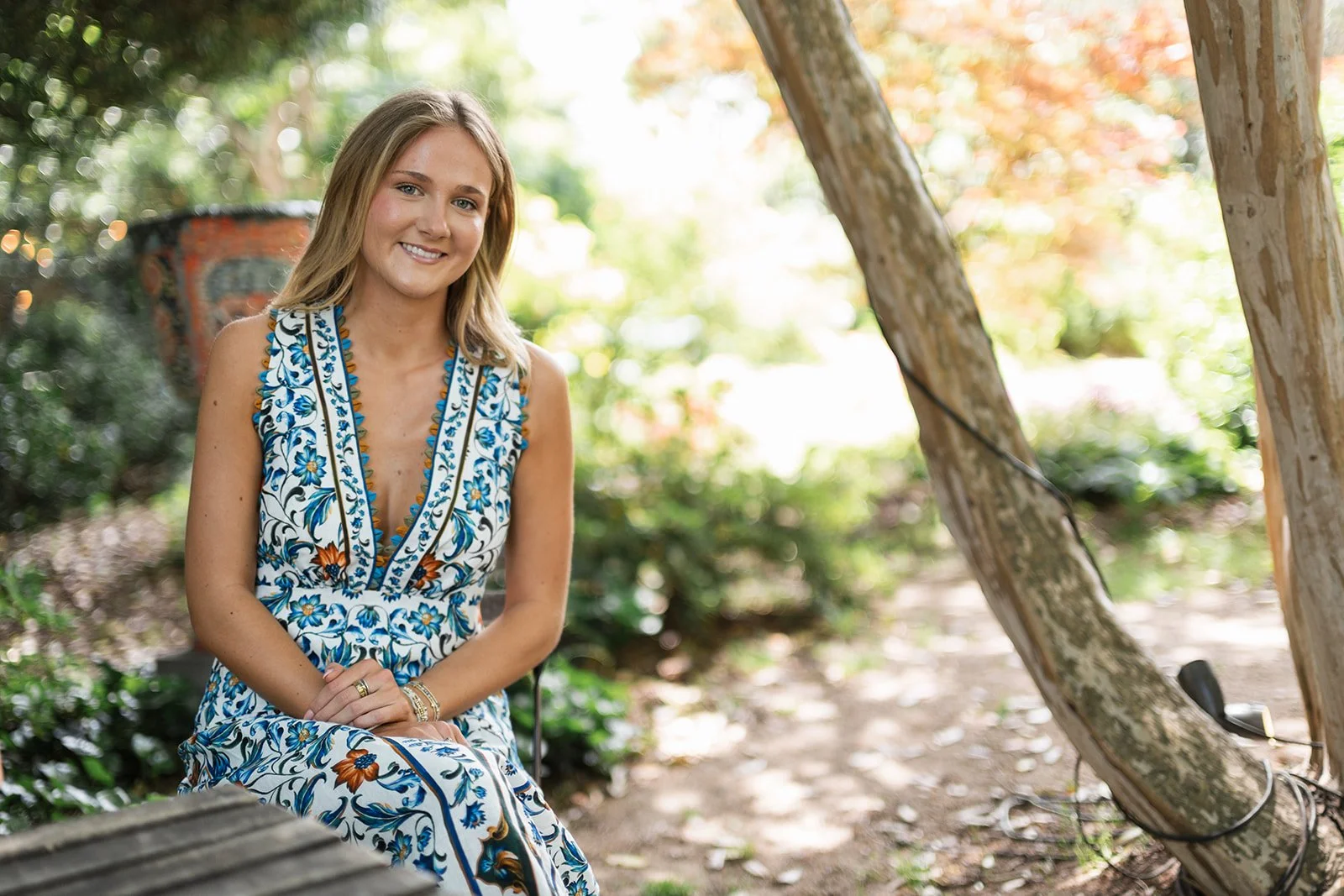 Young woman with blonde hair sitting outdoors at McGill Rose Garden in Charlotte, wearing a sleeveless dress with a floral pattern, smiling at the camera.